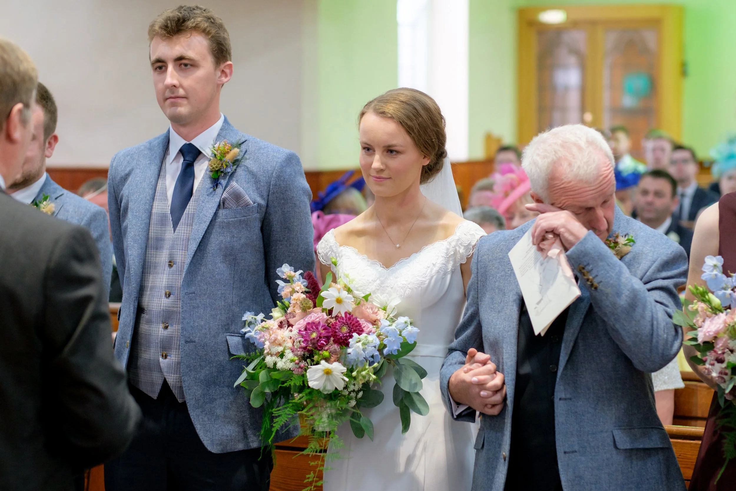 A bride holding a bouquet of pink, white, and blue flowers stands next to a groom in a gray suit during a wedding ceremony inside a church. An older man next to her appears emotional, wiping his eyes.