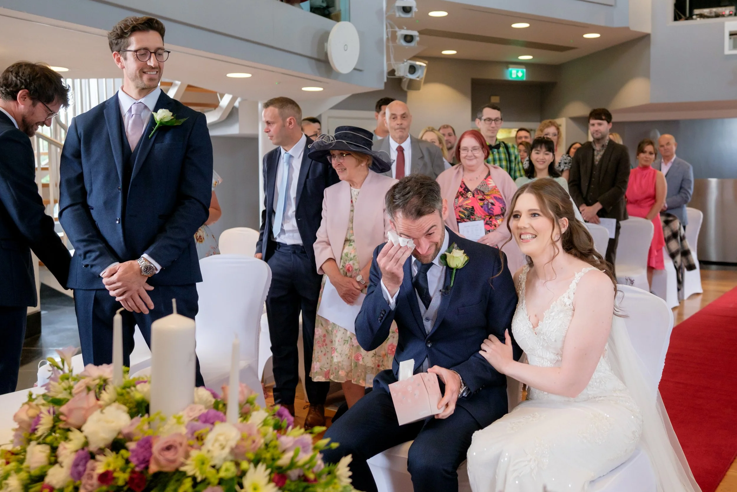 A wedding ceremony with brides and grooms, family, and friends in a modern indoor venue. The bride and groom are seated, smiling and the groom is wiping tears, holding a gift box. Guests are standing and watching, with some smiling warmly.