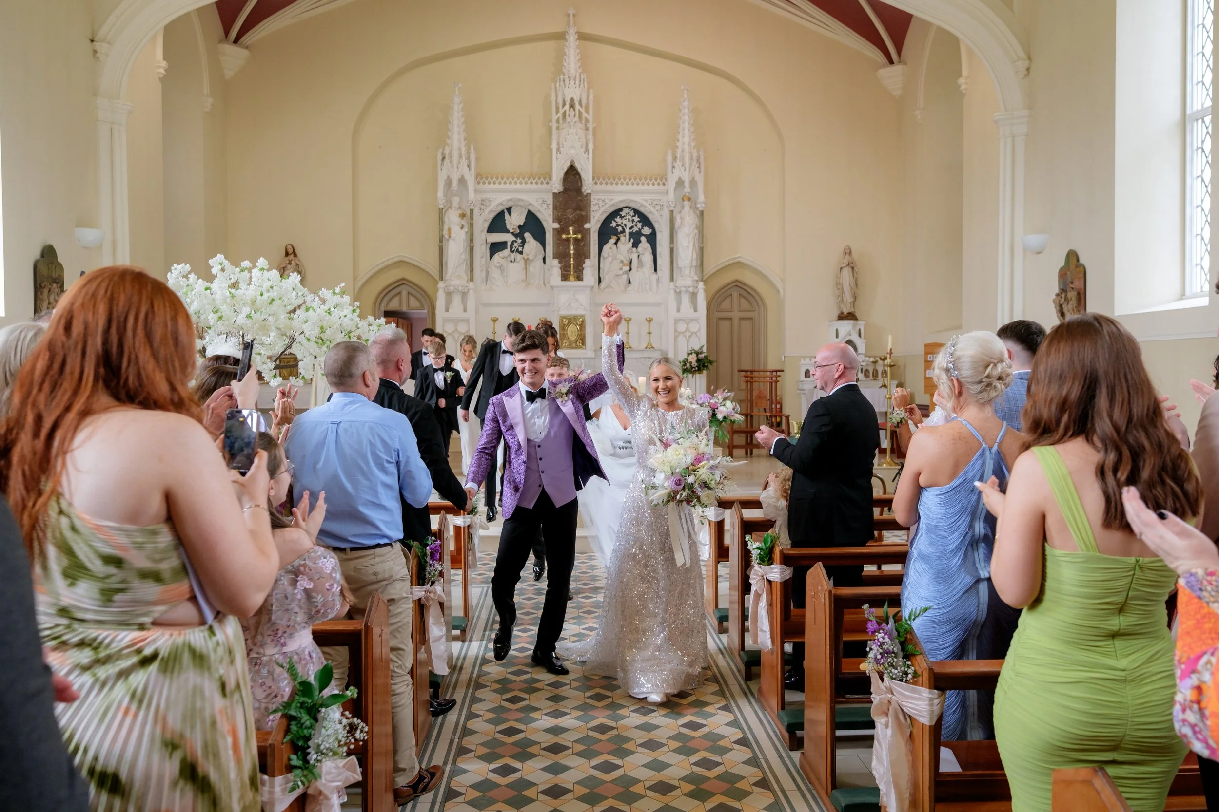 A newlywed couple walking down the aisle in a church, smiling and holding hands, with guests clapping and celebrating around them.