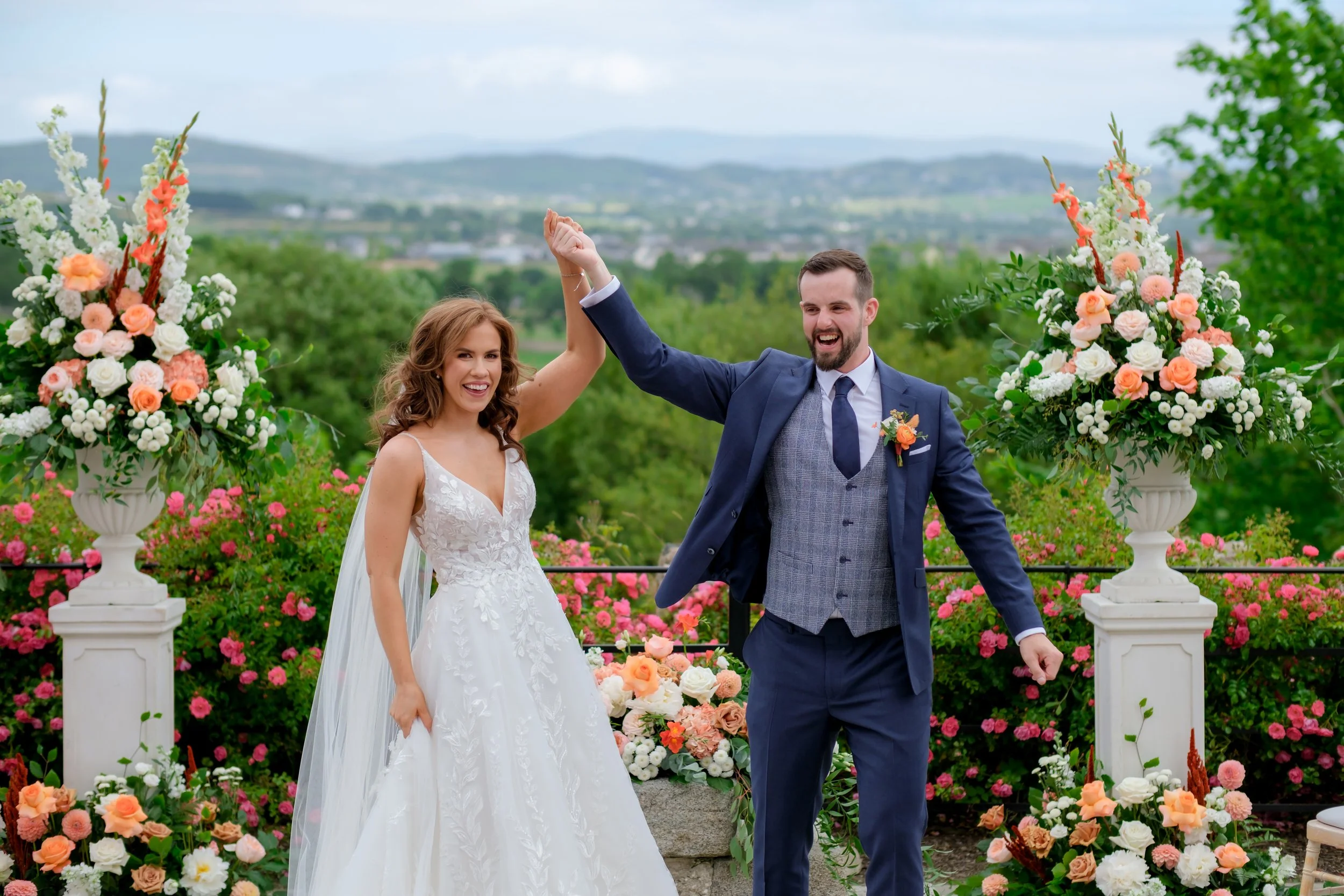 A newlywed couple celebrating outdoors on their wedding day, with the woman in a white lace wedding gown and the man in a blue suit, holding hands and smiling amid large floral arrangements and a scenic landscape.