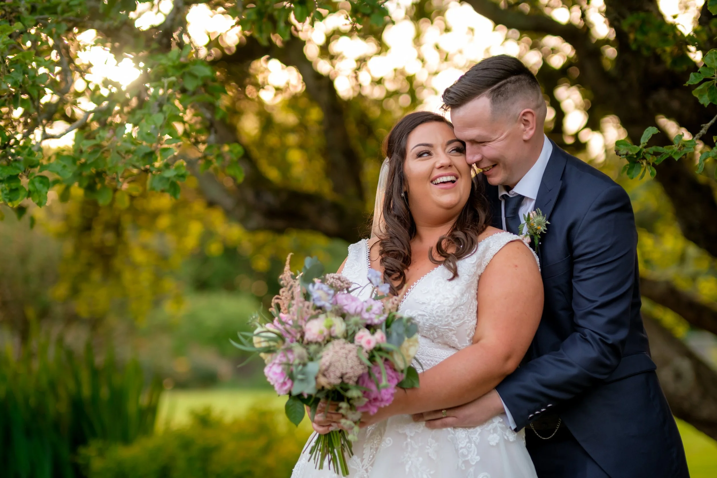 A happy bride and groom embrace outdoors during sunset, with the bride holding a bouquet of pink, purple, and white flowers, under a large tree with green leaves.