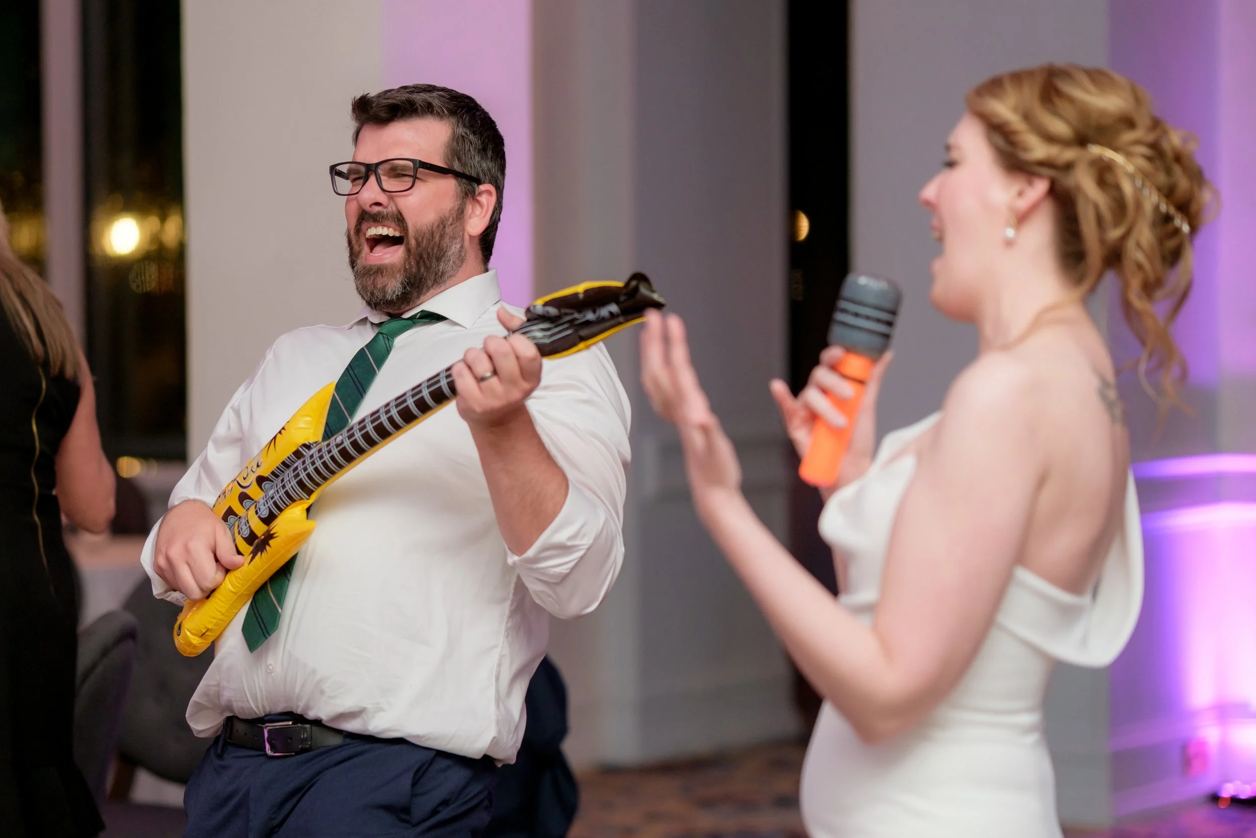 A man wearing glasses, a white shirt, a green striped tie, and navy pants is singing and playing a toy guitar. A woman with red hair styled in curls, wearing a white off-shoulder dress, is holding a microphone and singing back to him at an indoor par