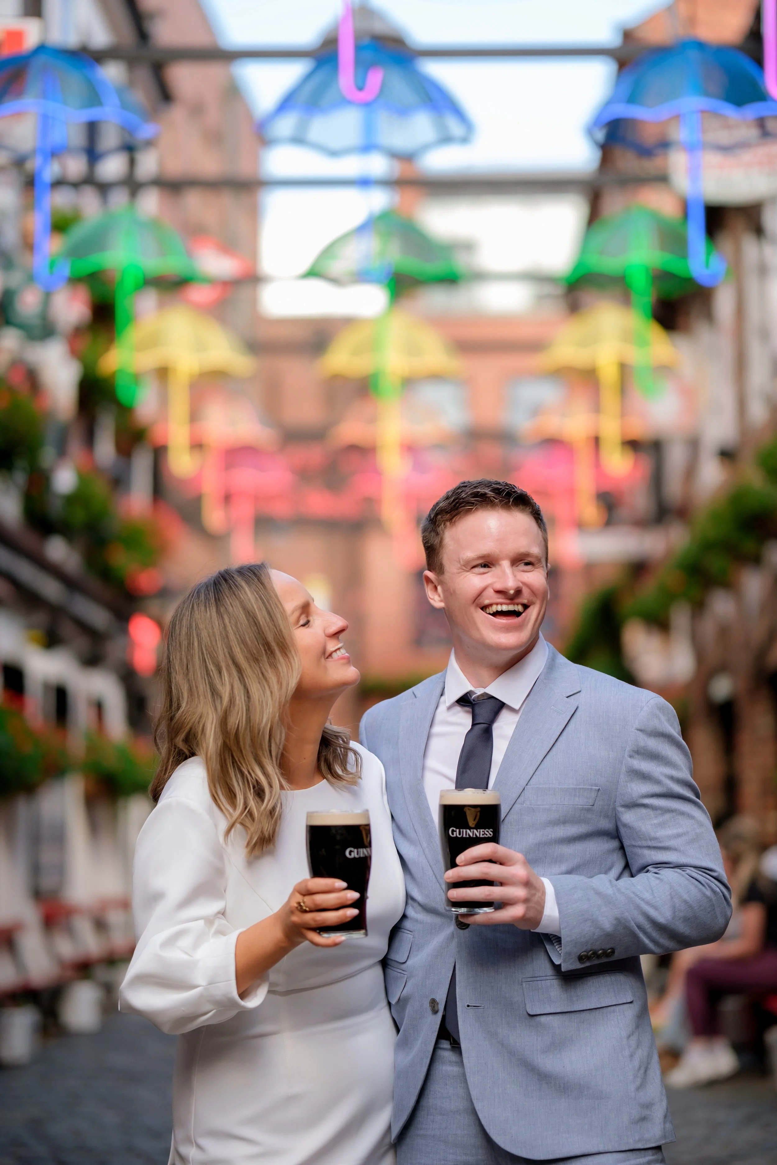 A man and woman dressed in formal attire holding pints of Guinness beer and smiling at each other in an outdoor pub or street setting decorated with colorful umbrellas hanging above.