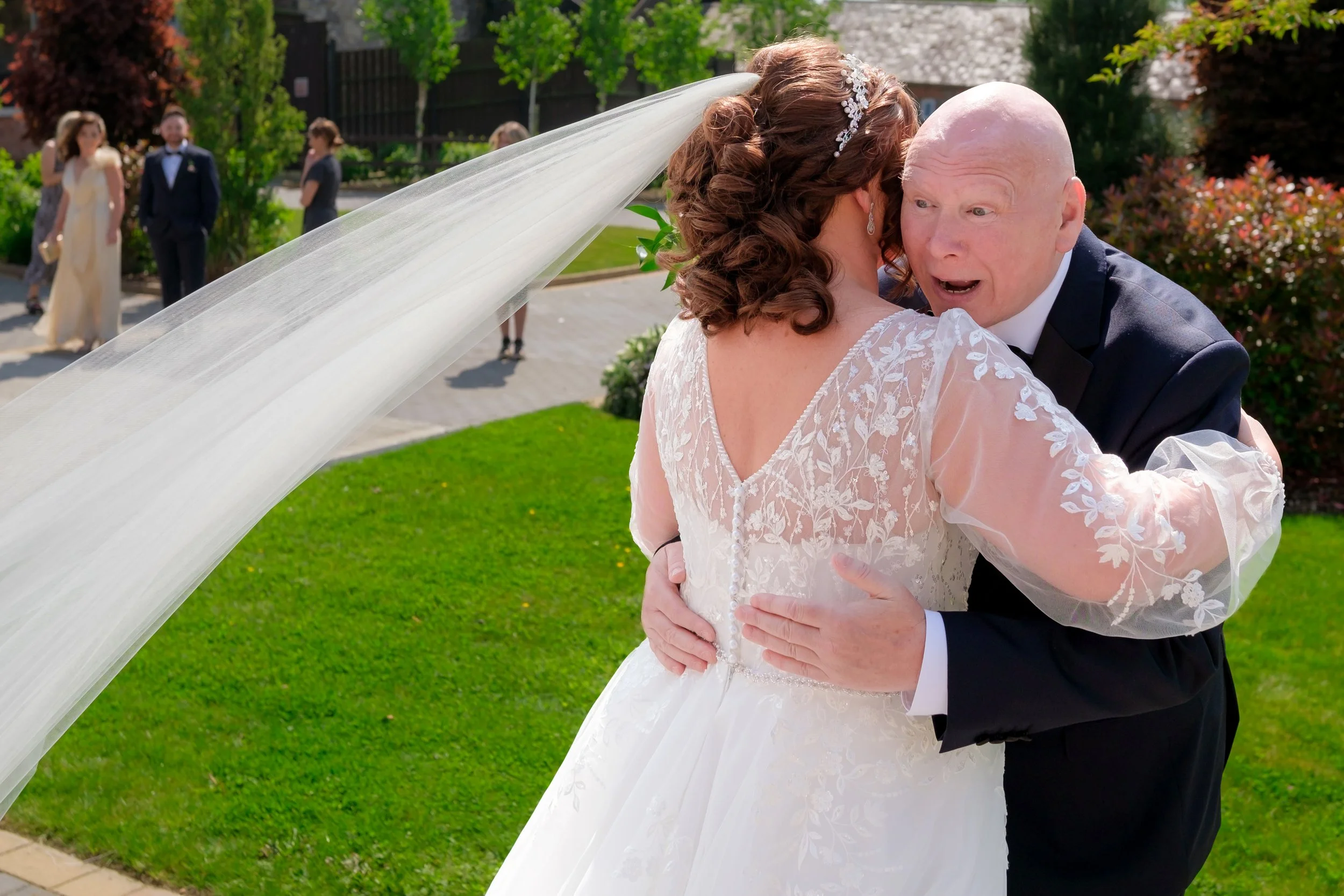 A bride and an older man, possibly her father, sharing a joyful embrace outdoors on her wedding day, with a wedding gown and veil, and other people in the background.