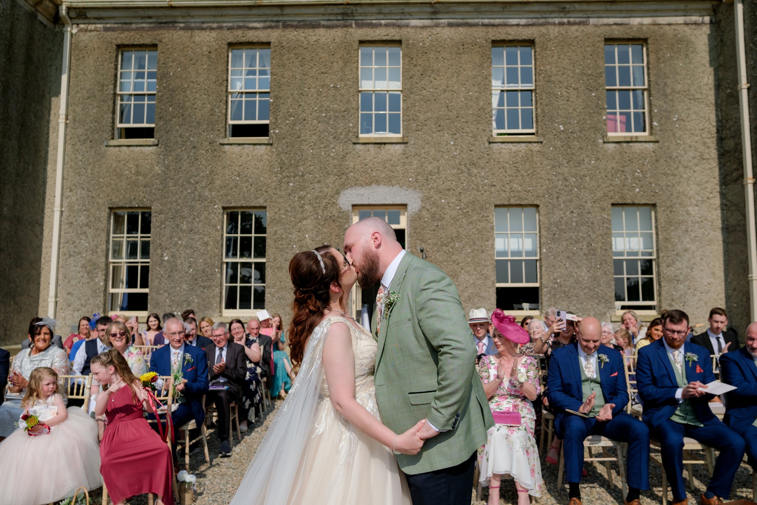 Bride and groom kissing during wedding ceremony outside with seated guests clapping and smiling.