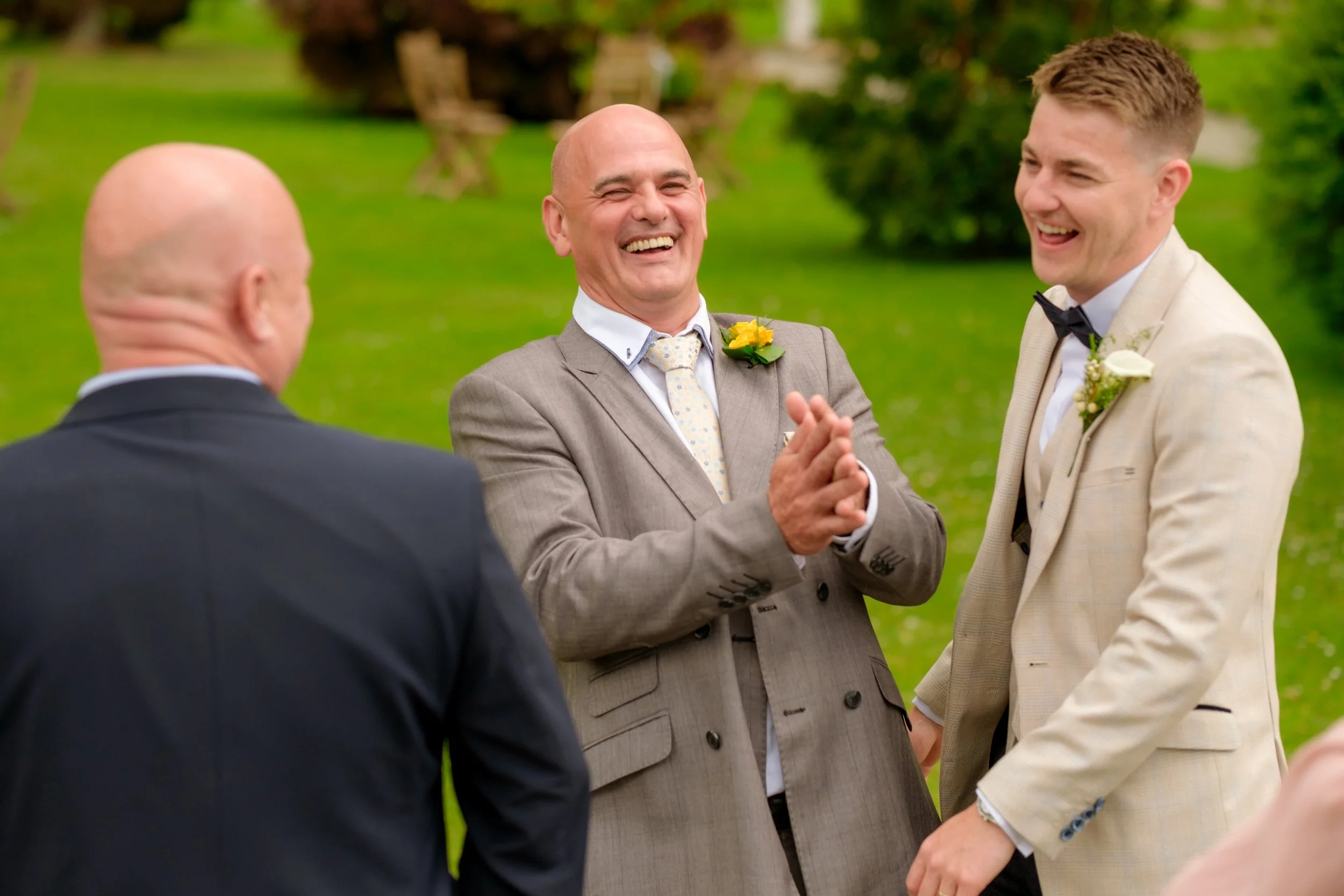 A group of men smiling and laughing outdoors during a wedding ceremony on a green lawn.
