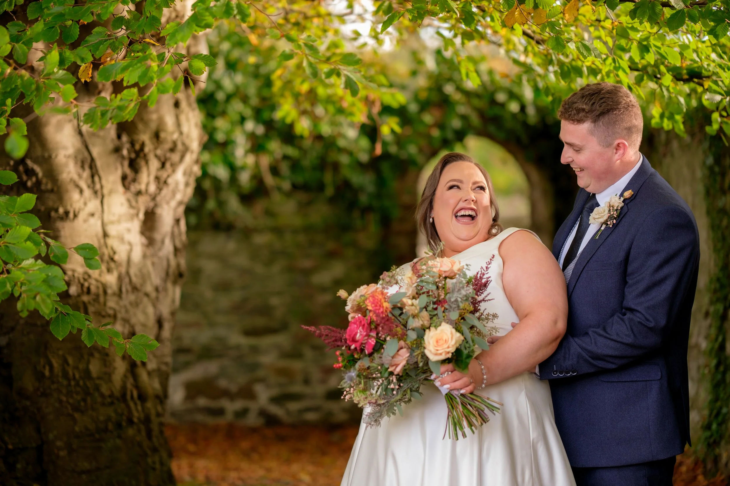 A happy bride in a white wedding dress holding a bouquet of pink and peach flowers, laughing with a smiling groom in a navy suit in a lush green outdoor setting with trees.