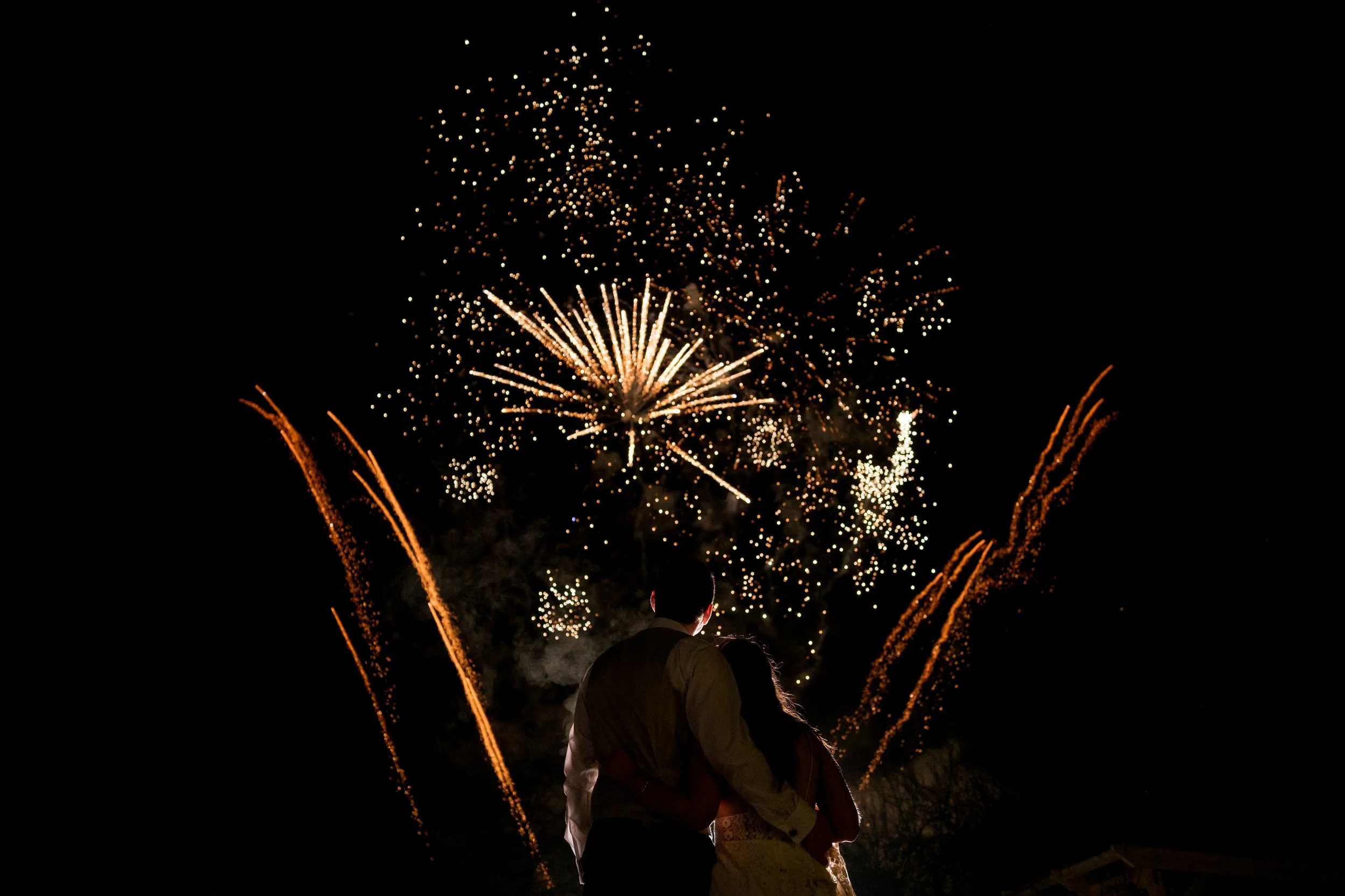 A couple stands close together watching fireworks light up the night sky.