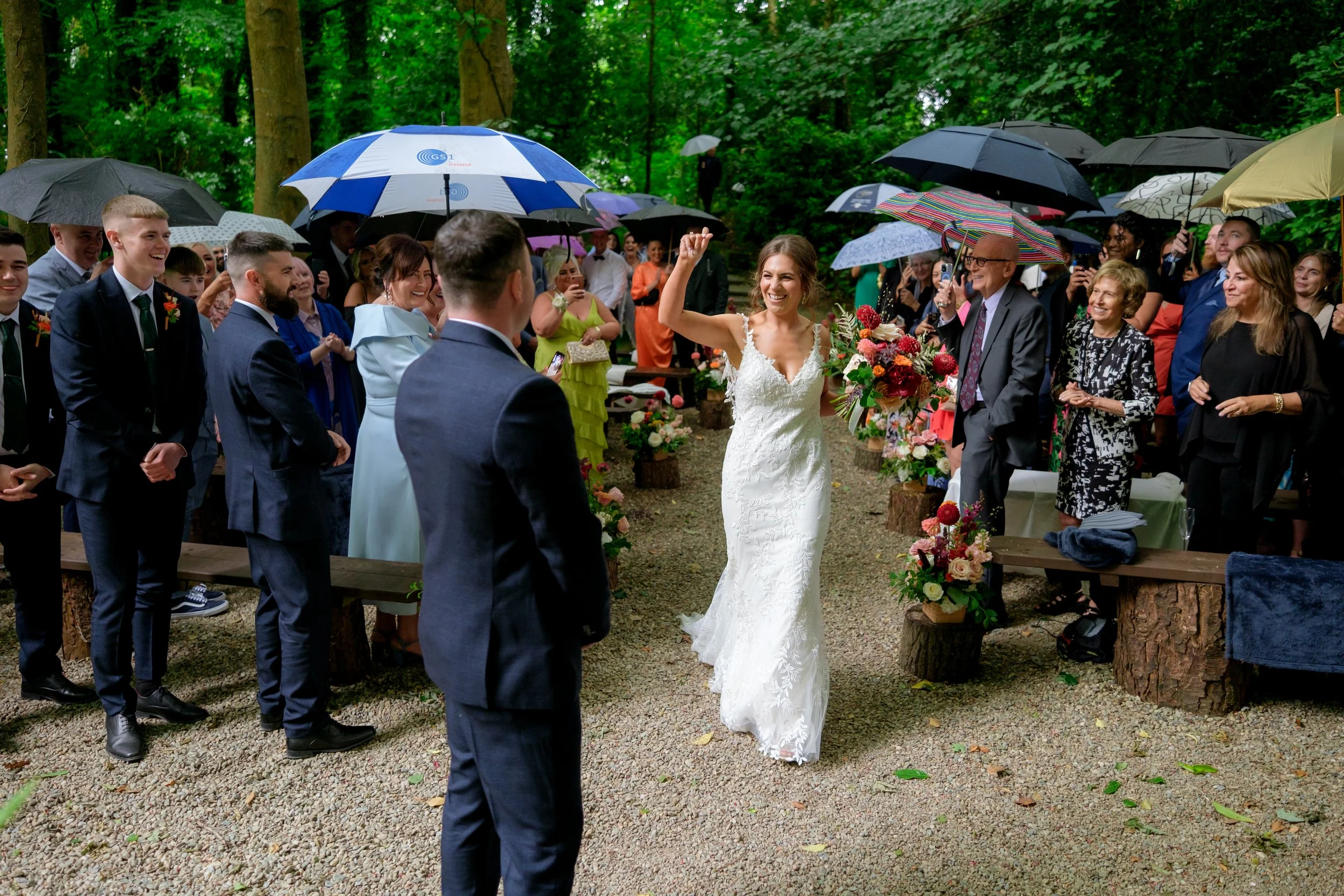 Bride walking down the aisle at an outdoor wedding reception, smiling and holding a bouquet, surrounded by guests holding umbrellas in a wooded area.