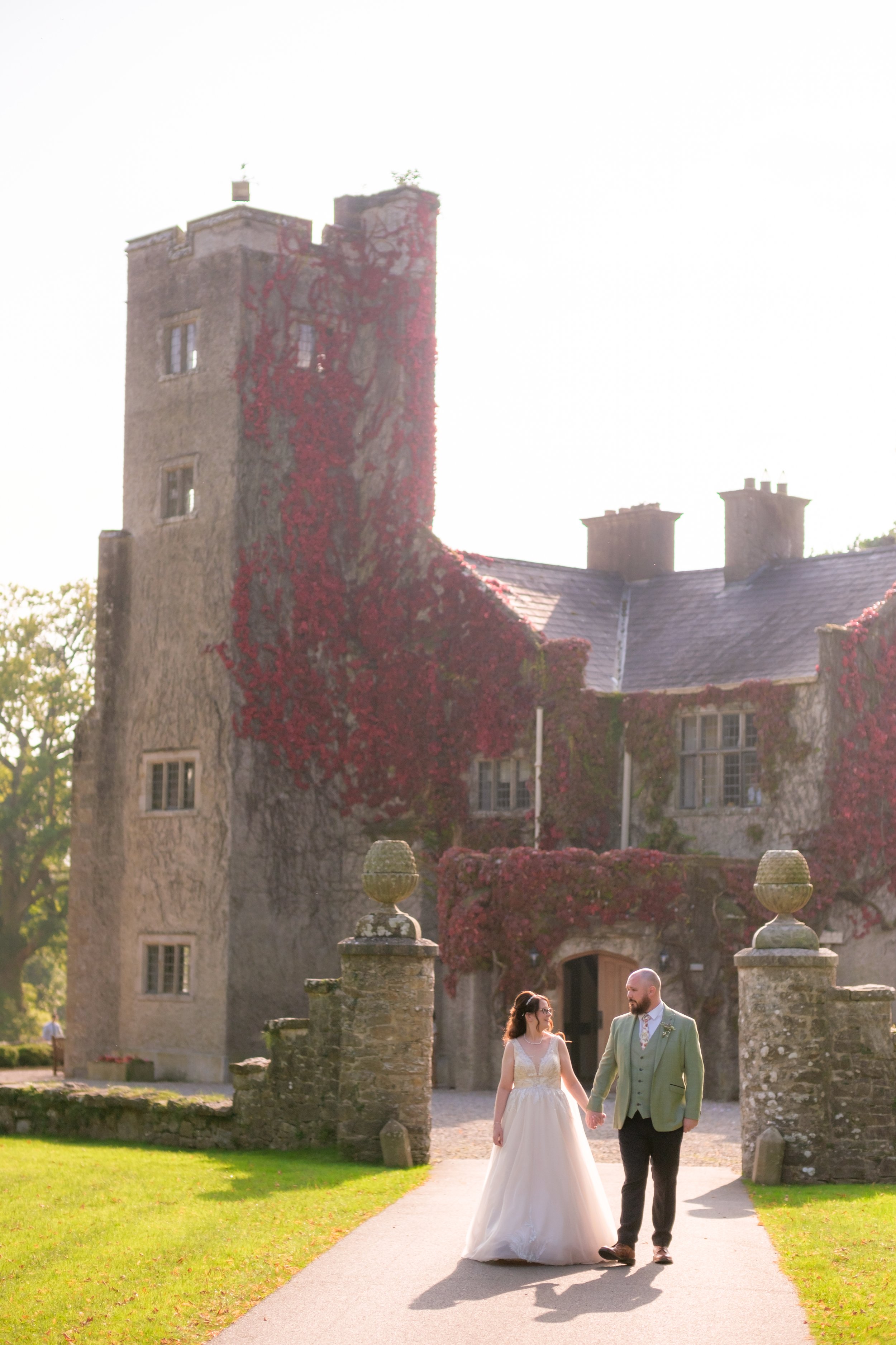 A bride and groom holding hands walk towards a historic castle with ivy-covered stone walls, under a bright, sunny sky.