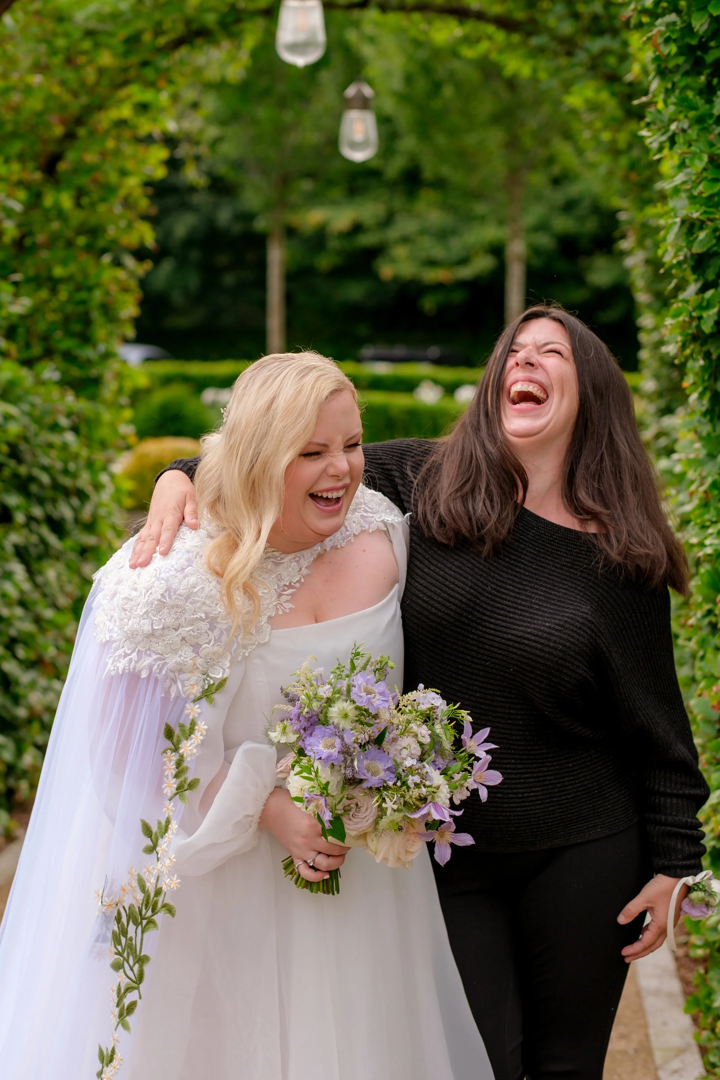 Two women laughing and embracing in a garden, one in a wedding dress holding a bouquet of flowers.