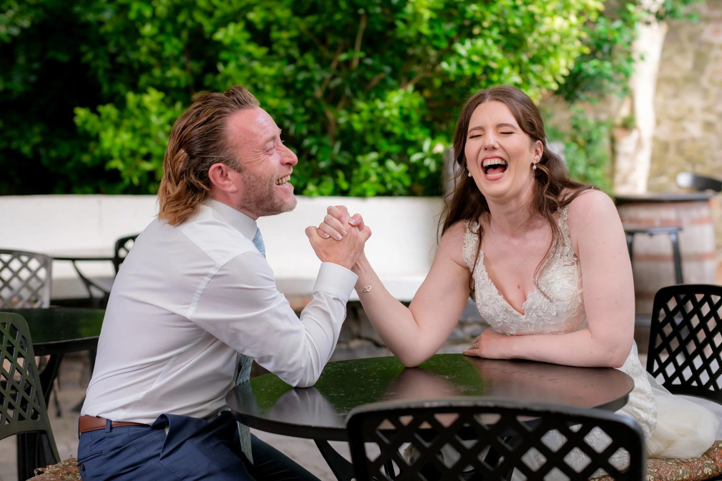 A man and a woman are sitting at a round table outdoors, smiling and laughing while arm wrestling. The woman is wearing a wedding dress and the man is dressed in formal attire. They are enjoying a joyful moment with greenery and outdoor furniture in 