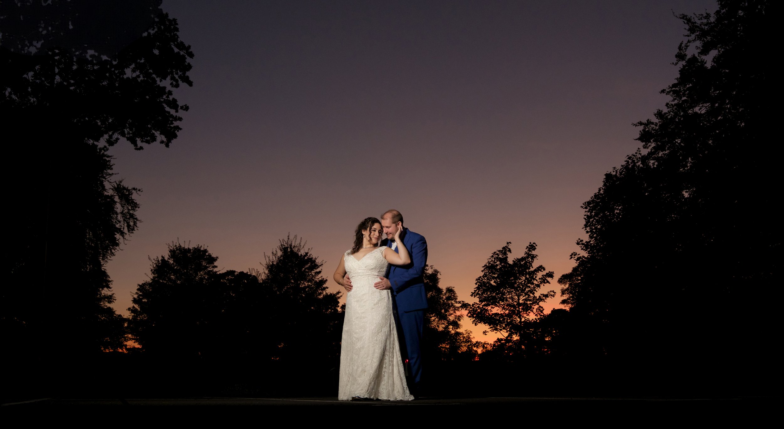 Bride and groom embracing at sunset with silhouetted trees in the background.