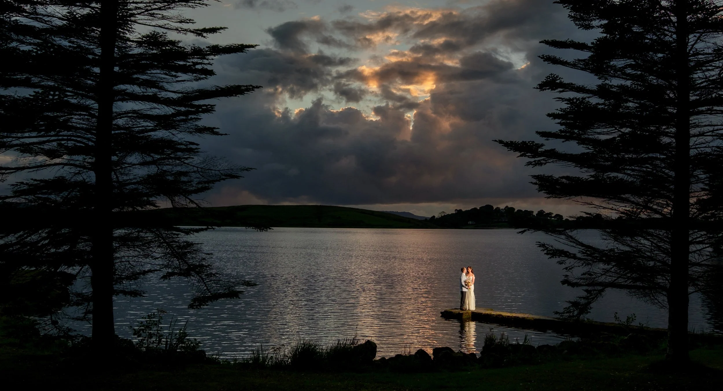 A couple in wedding attire standing on a small pier by a lake during sunset, with dark clouds and trees framing the scene.