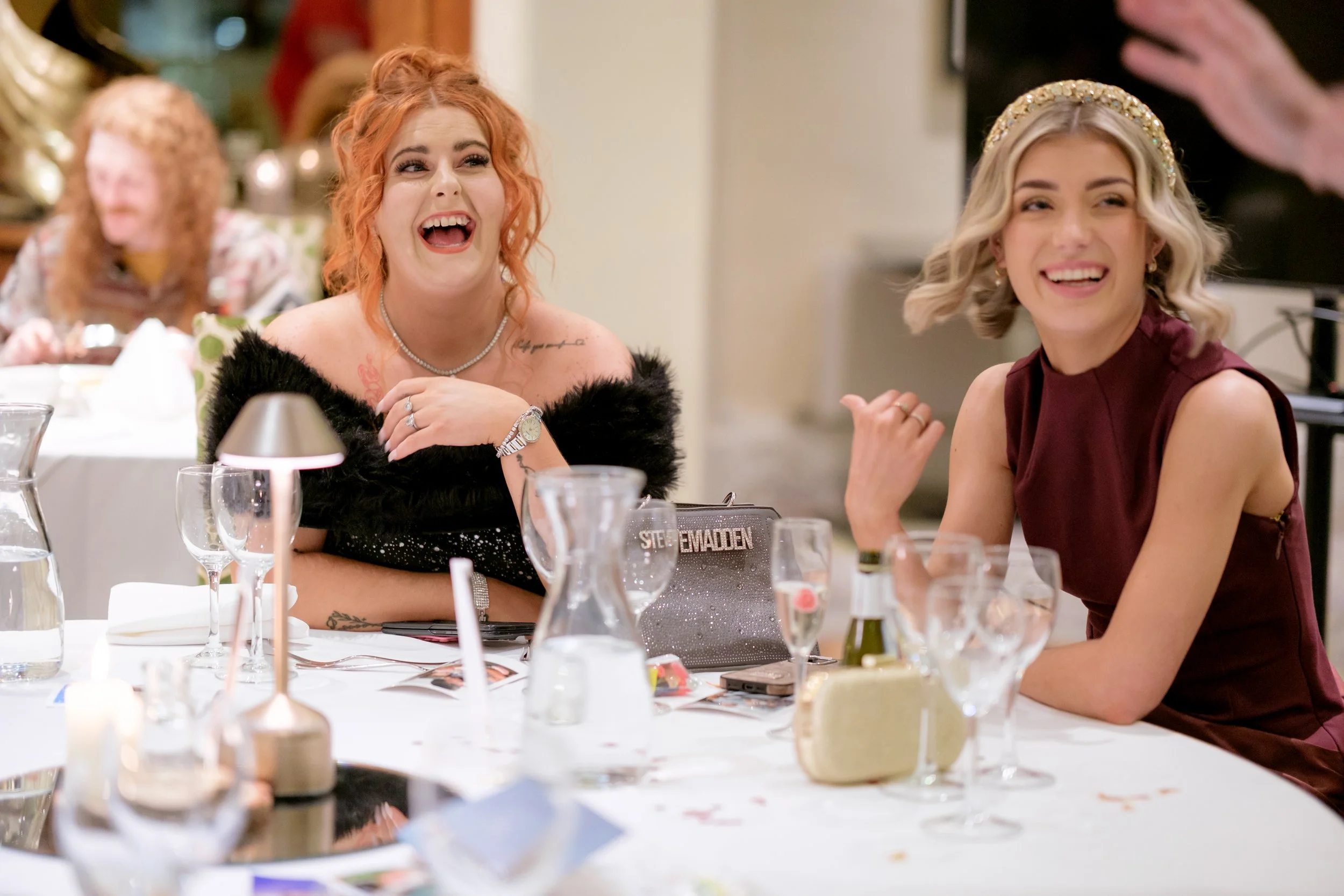 Two women laughing and enjoying themselves at a dinner party or celebration. One has red hair and is wearing a black fuzzy off-shoulder top, the other has blonde hair and is wearing a sleeveless dark red dress with a glittery headband. They are seate