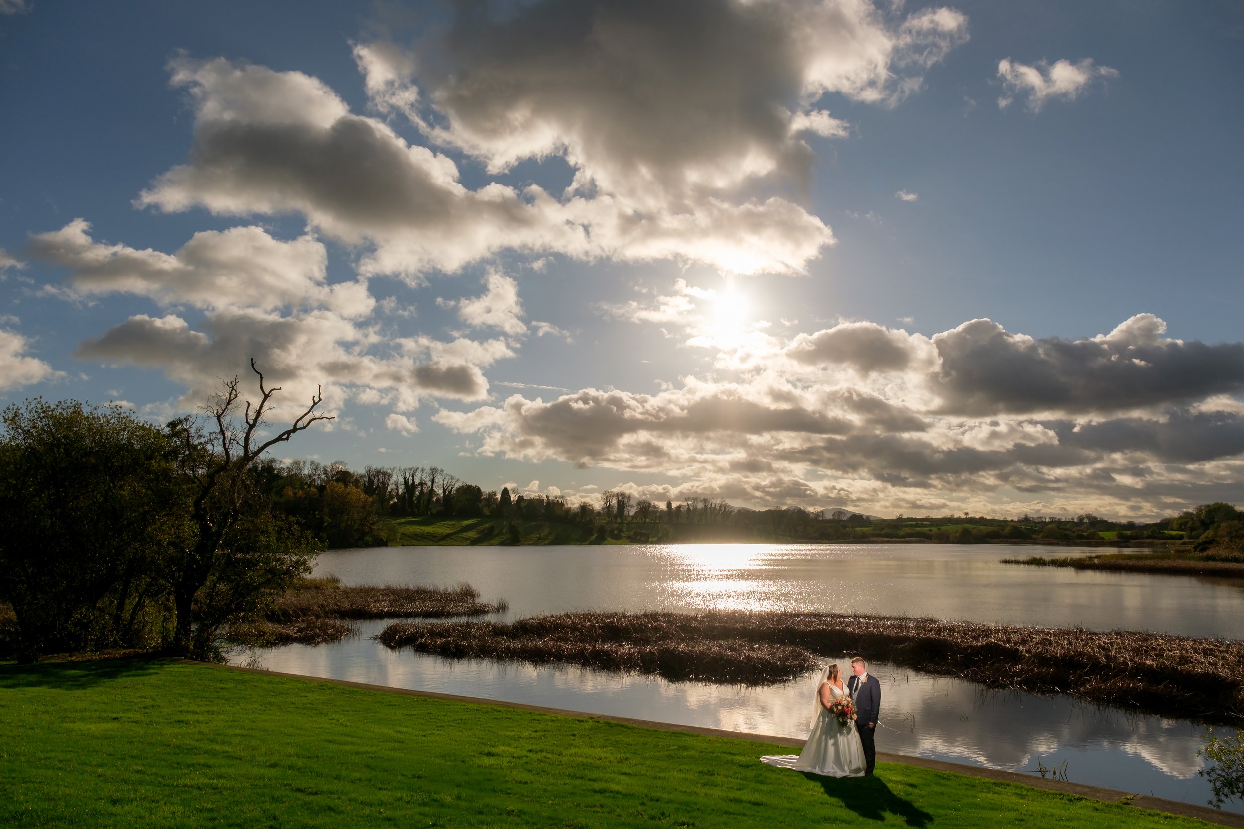 A bride and groom standing on a grassy area near a lake, with trees, water, and a partly cloudy sky in the background during sunset.