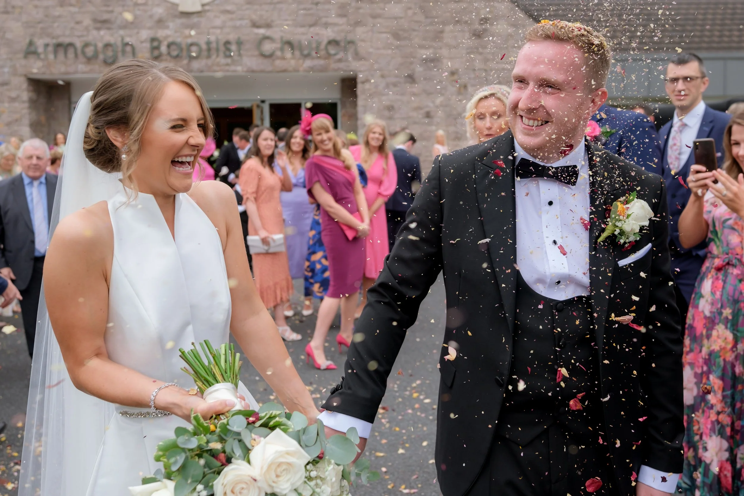 Bride and groom smiling and holding hands amidst confetti at their wedding celebration outside a church, with wedding guests in colorful attire in the background.