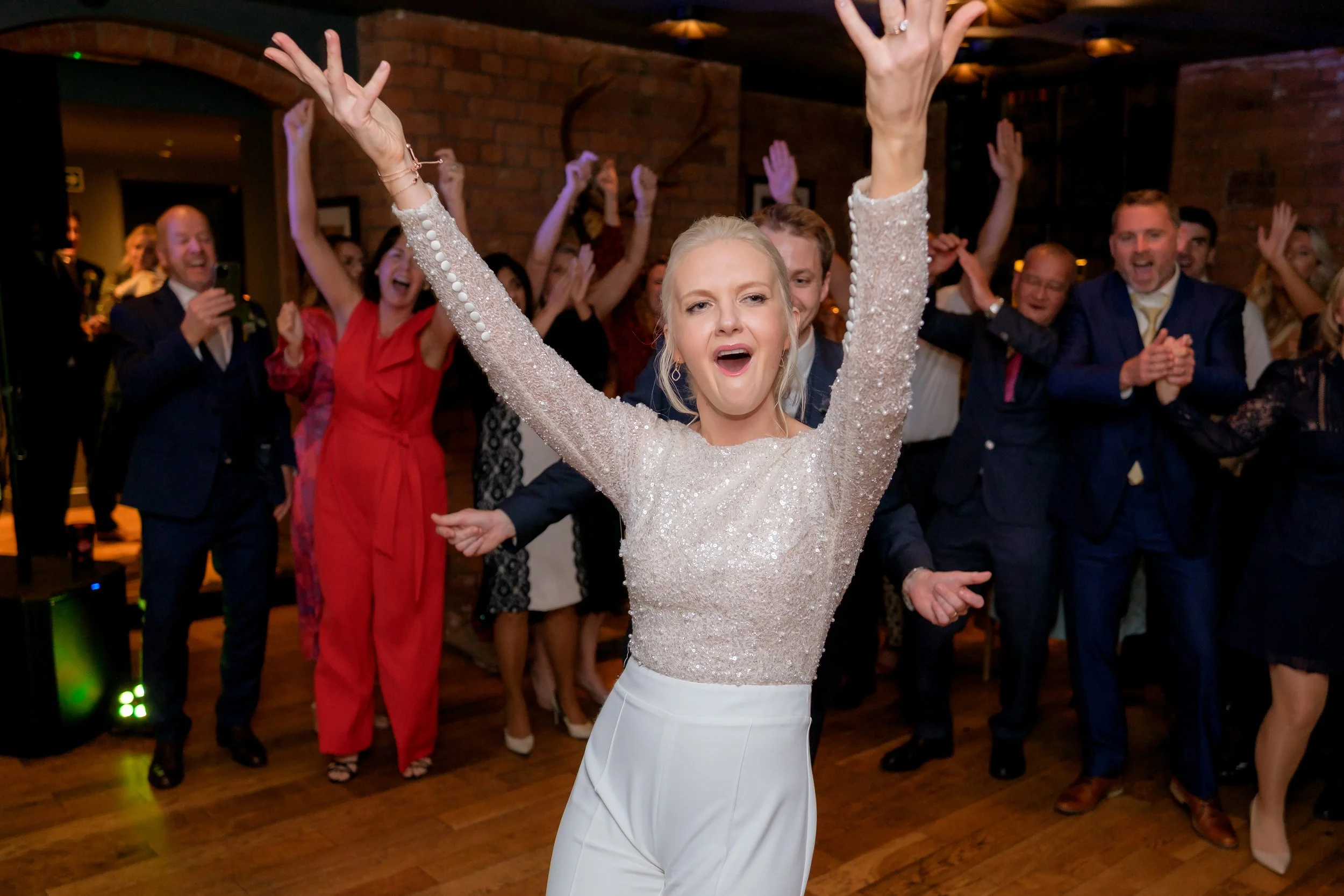 Woman in a sparkling long-sleeve top and white pants dancing with arms raised, surrounded by smiling people in formal attire at a celebration or wedding reception.