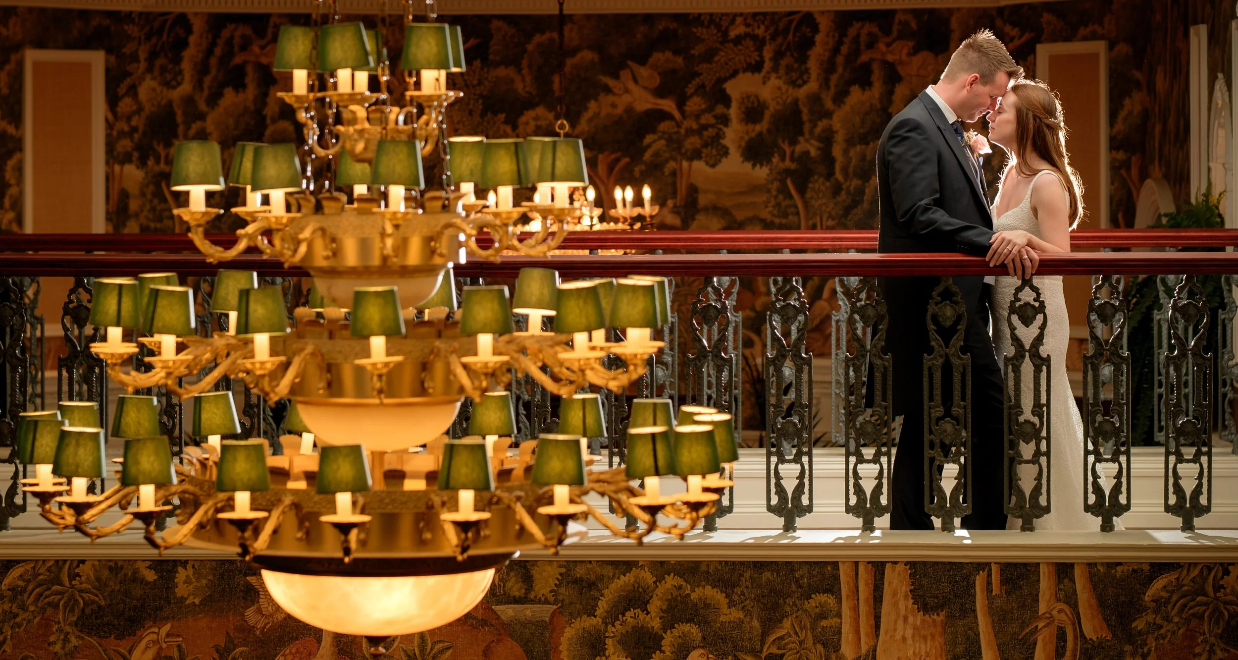 A couple dressed in wedding attire standing close on a balcony, touching foreheads and holding hands, with large green-shaded chandeliers hanging above them.