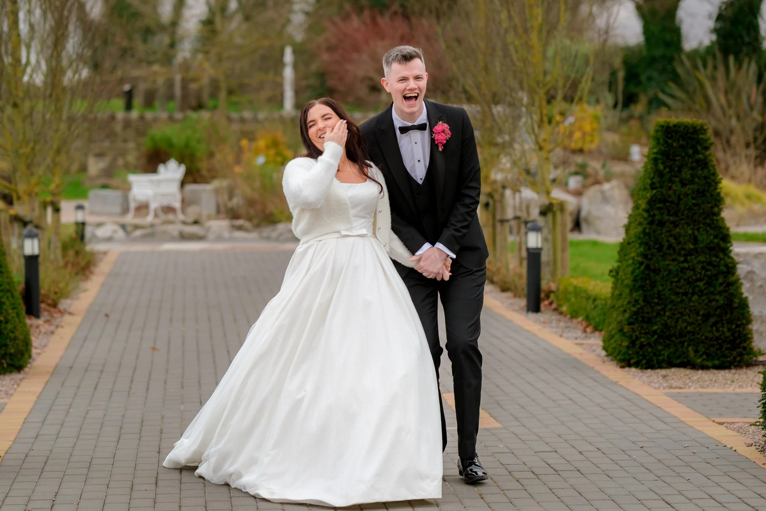 A newlywed couple in wedding attire, the bride in a white gown and the groom in a black tuxedo, laughing and holding hands outdoors on a paved pathway lined with bushes and trees.
