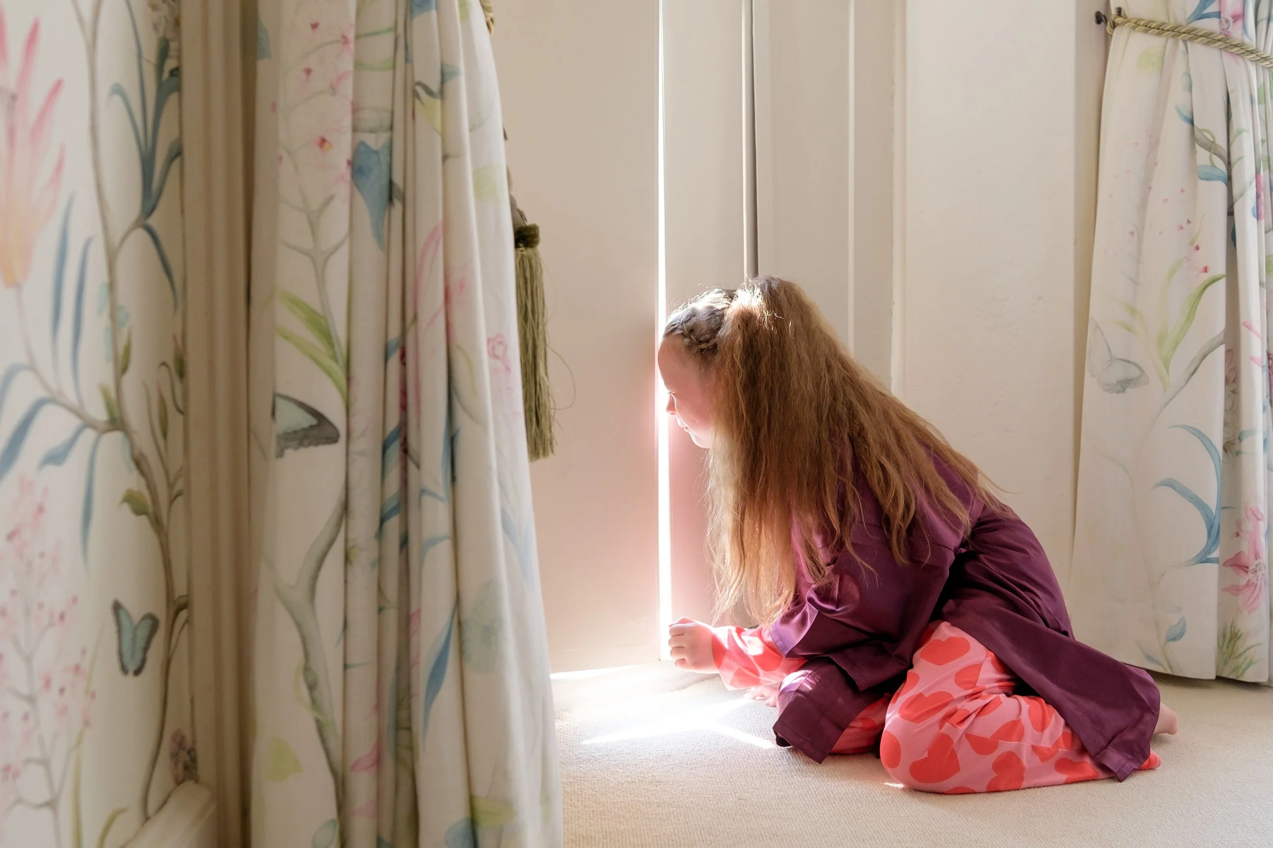 A young girl with long, curly hair kneeling on the carpet, looking out through a partially opened door as sunlight streams in.