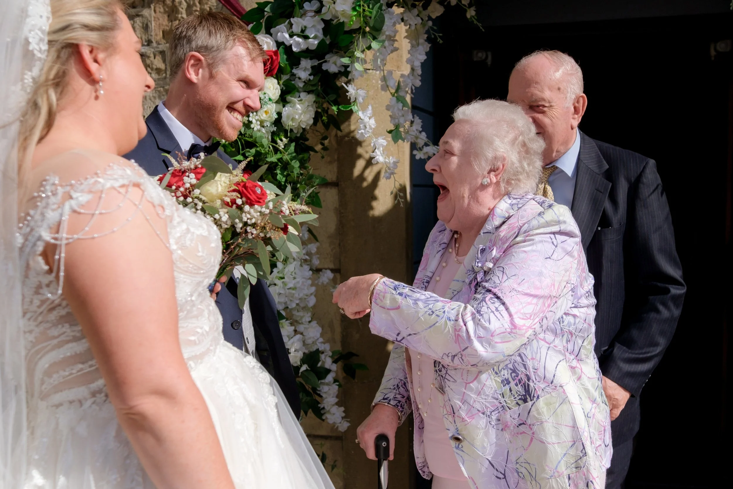 A joyful bride and groom smiling at an elderly woman during a wedding celebration. The elderly woman, holding a cane, is laughing and pointing at the bride. A man and a woman stand behind her, observing the interaction. The background is decorated wi