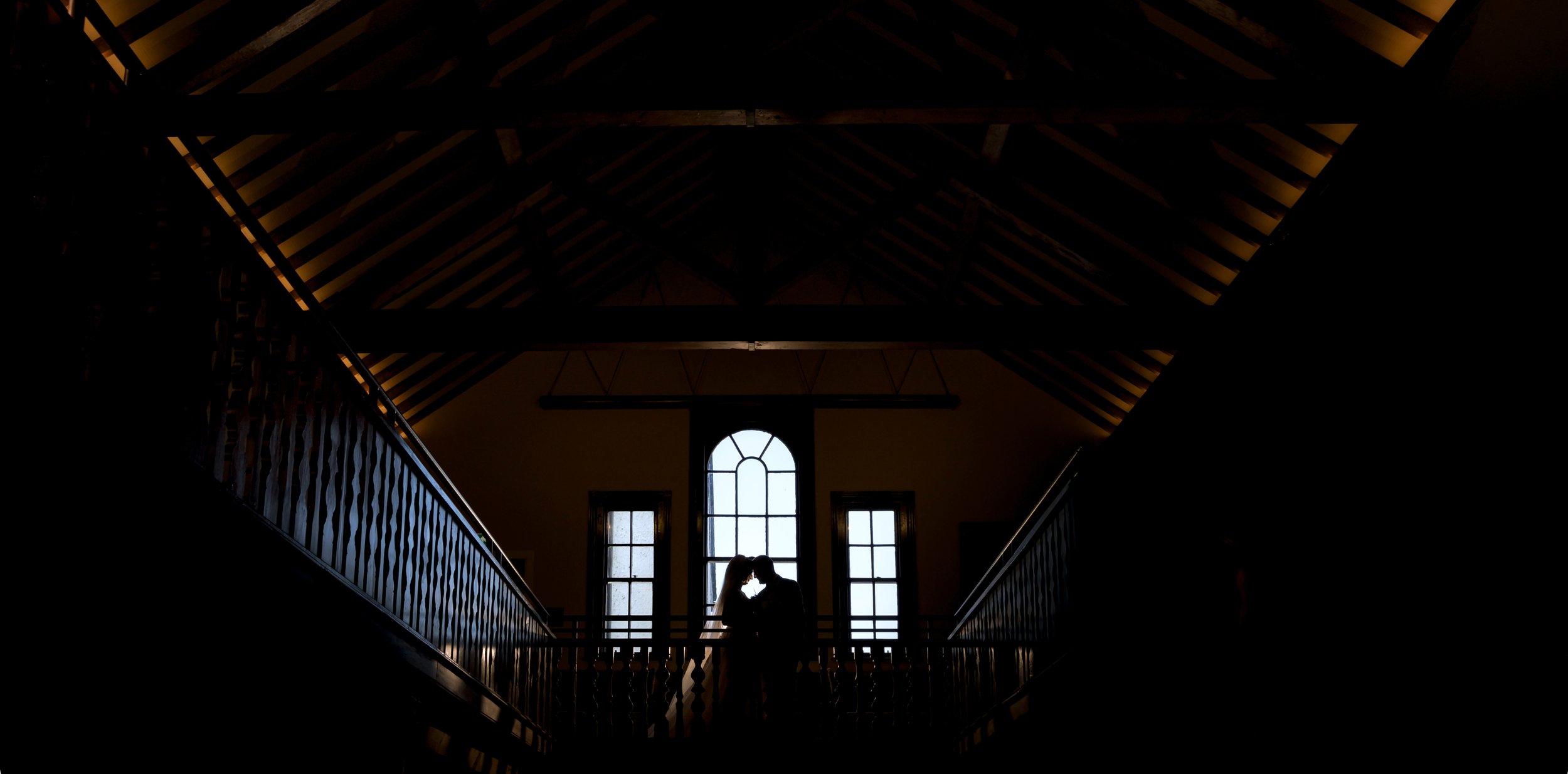 Silhouette of a couple kissing in front of large windows inside a dimly lit building with wooden beams on the ceiling and a balcony railing.