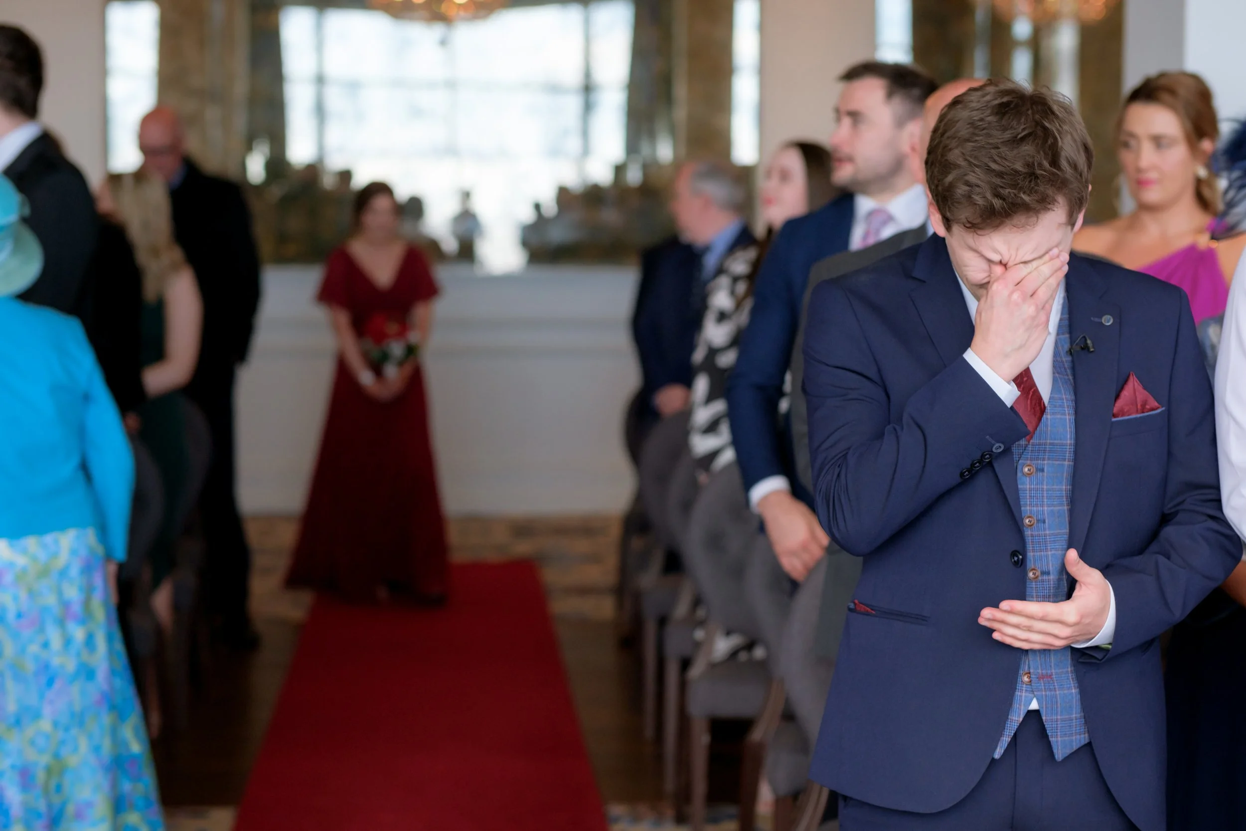 A young boy in a blue suit with a red tie is holding his face in his hands, appearing emotional, while people are standing and seated in the background at a formal event.