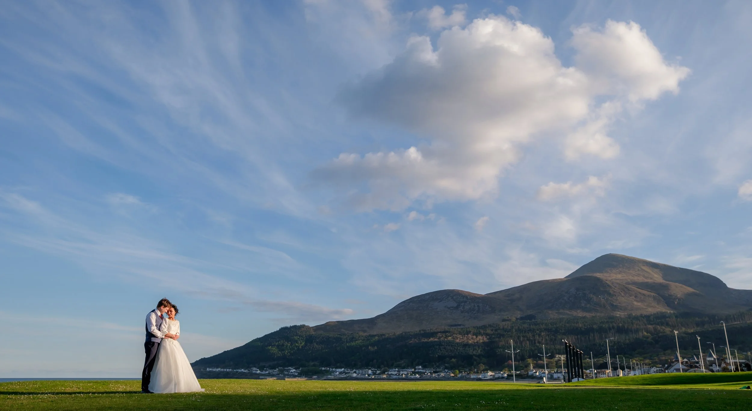 A bride and groom standing together on a grassy field with mountains in the background, under a partly cloudy sky.
