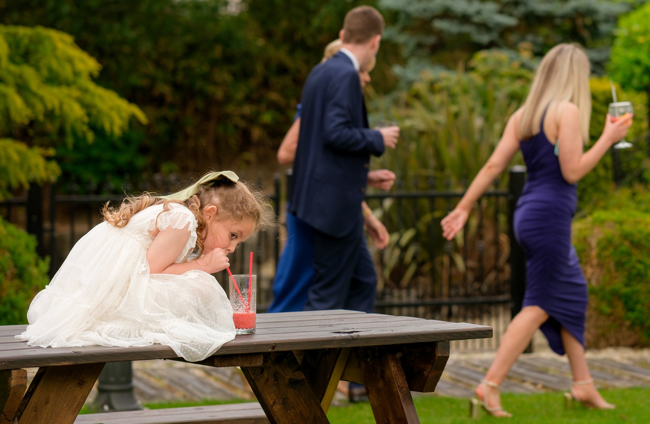 A young girl in a white dress with a green ribbon in her hair is sitting on a wooden picnic table, drinking a pink beverage with striped straws. In the background, two adults, a man in a dark suit and a woman in a purple dress, are walking and holdin