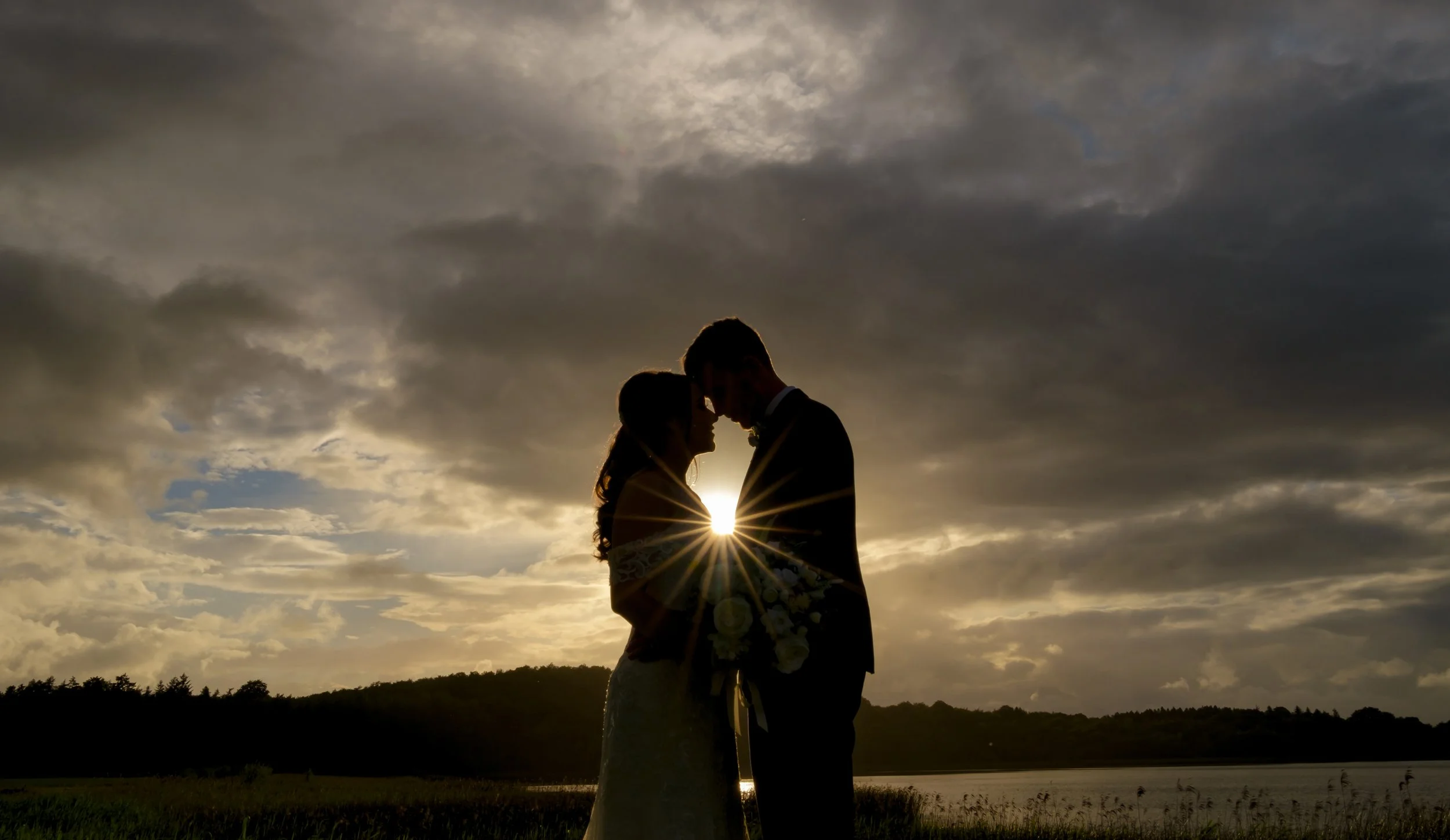Silhouette of a couple standing close together with their foreheads touching, holding a bouquet, during sunset near a river with a cloudy sky.