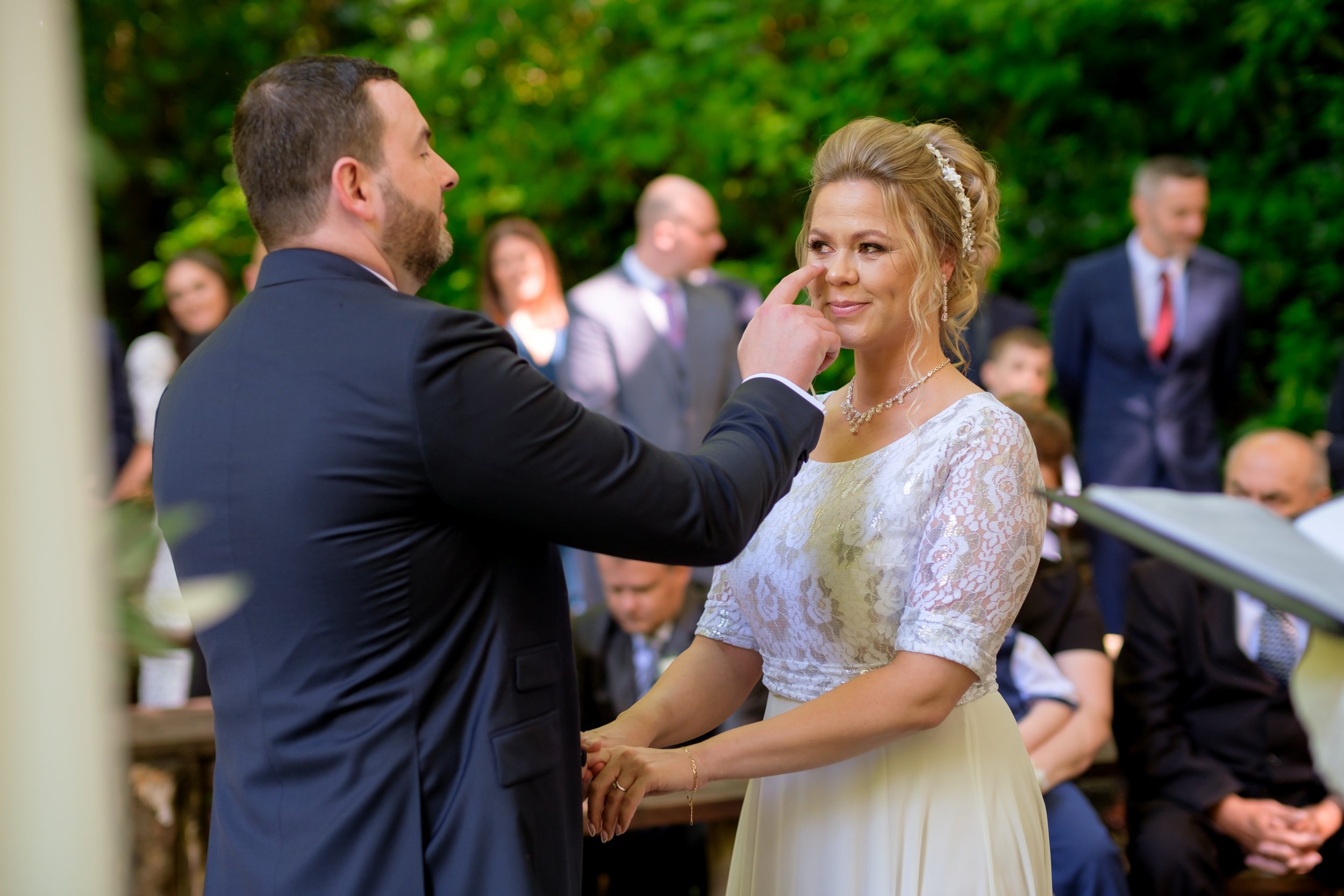 A bride and groom during their wedding ceremony outdoors, with the groom touching the bride's nose playfully as she smiles, surrounded by guests.