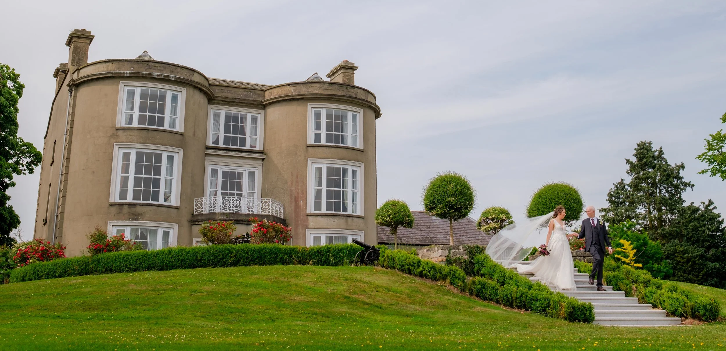 A bride and groom descending outdoor stairs near a large, historic mansion with multiple windows, surrounded by manicured lawns and trees.