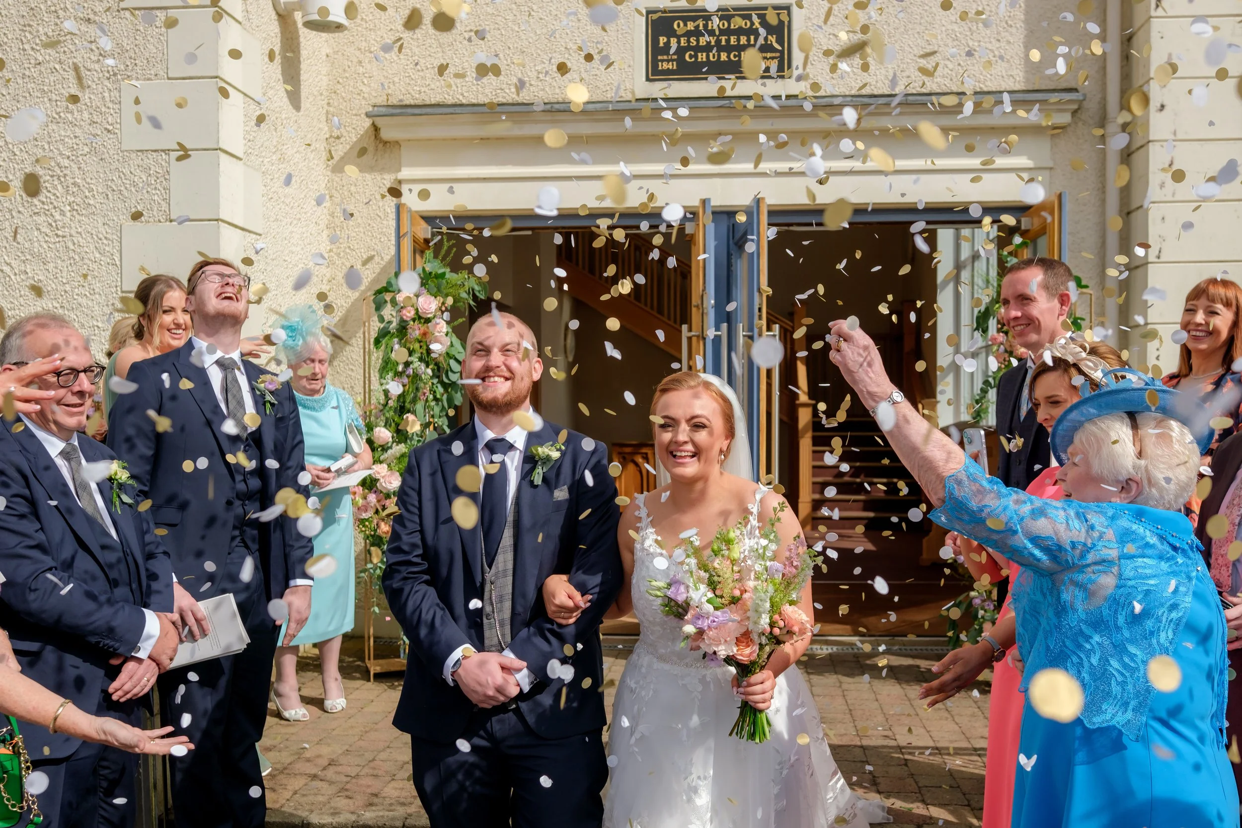 A happy bride and groom walking out of a church surrounded by friends and family, with confetti falling around them during their wedding celebration.