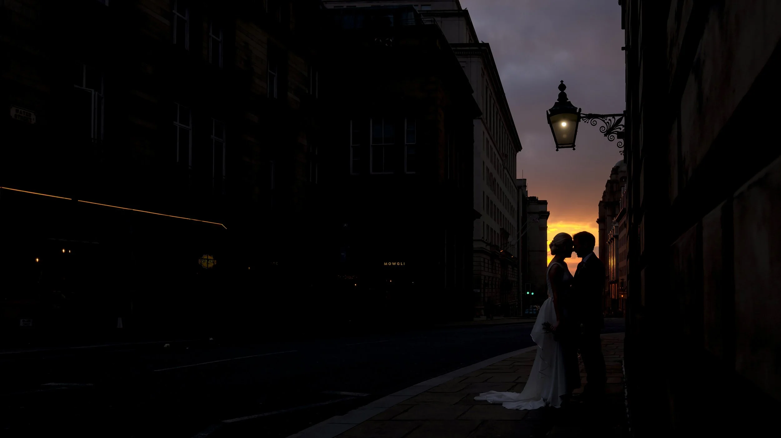 A silhouette of a couple in wedding attire standing close together on a city street at sunset, with buildings and a street lamp visible in the background.