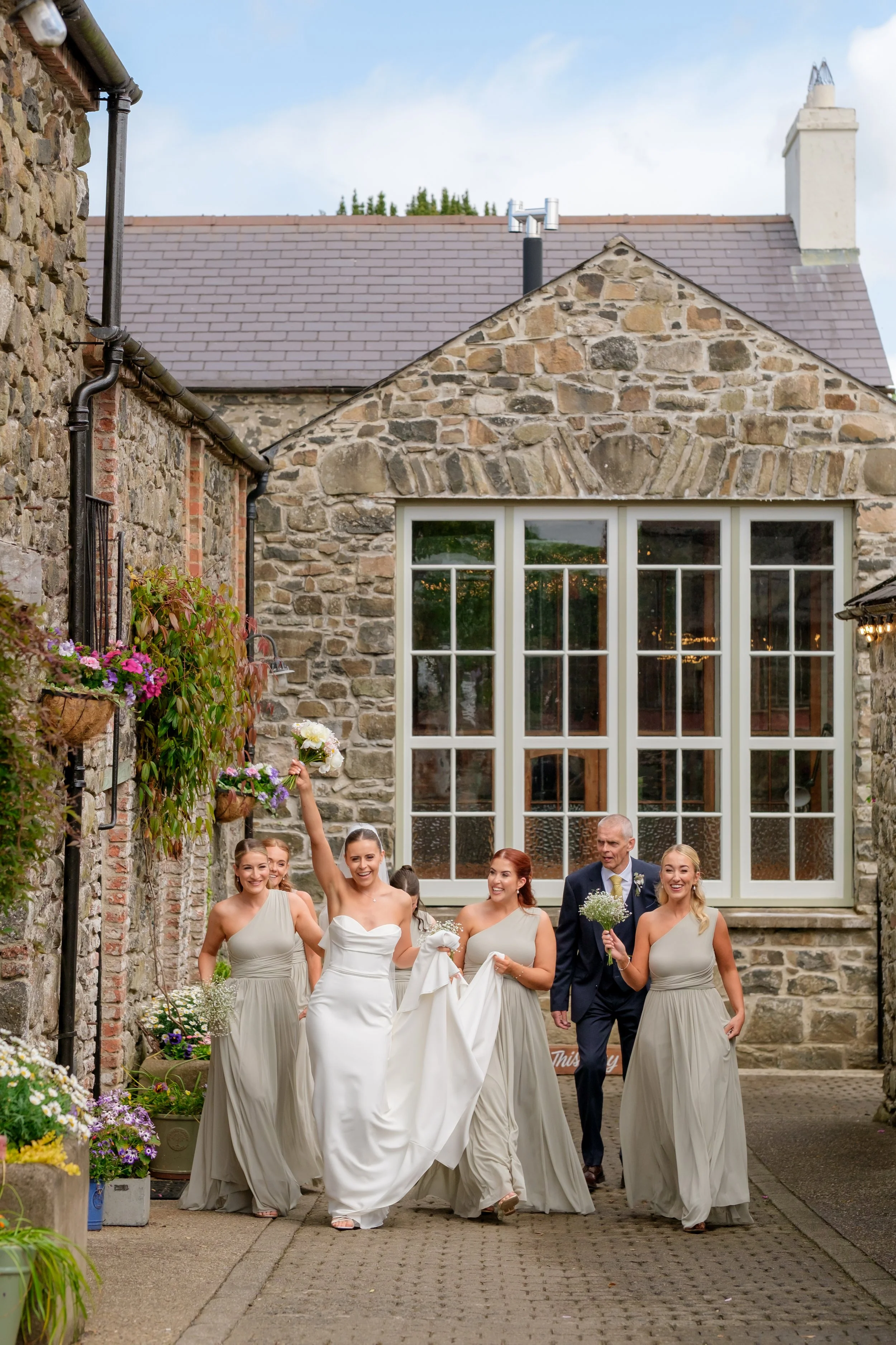 A bride in a white wedding dress and five bridesmaids in matching grey dresses walking on a cobblestone path outside a stone building, celebrating and smiling. The bride is holding a bouquet and raising her hand, while the others hold flowers and the
