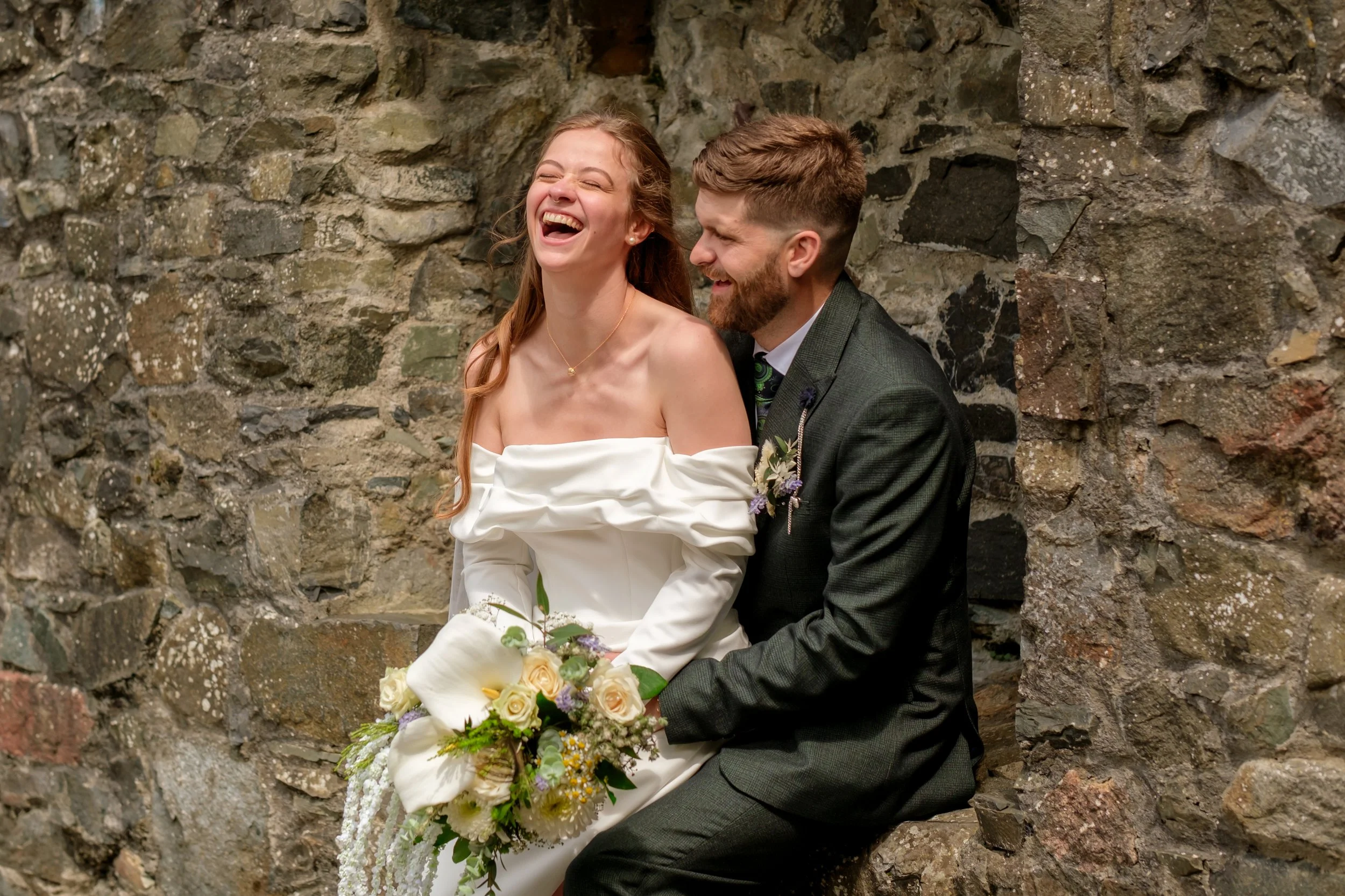 A bride and groom sitting together against a stone wall during their wedding, both smiling and enjoying the moment. The bride holds a bouquet and wears an off-the-shoulder white dress, while the groom is in a dark suit with a boutonniere.