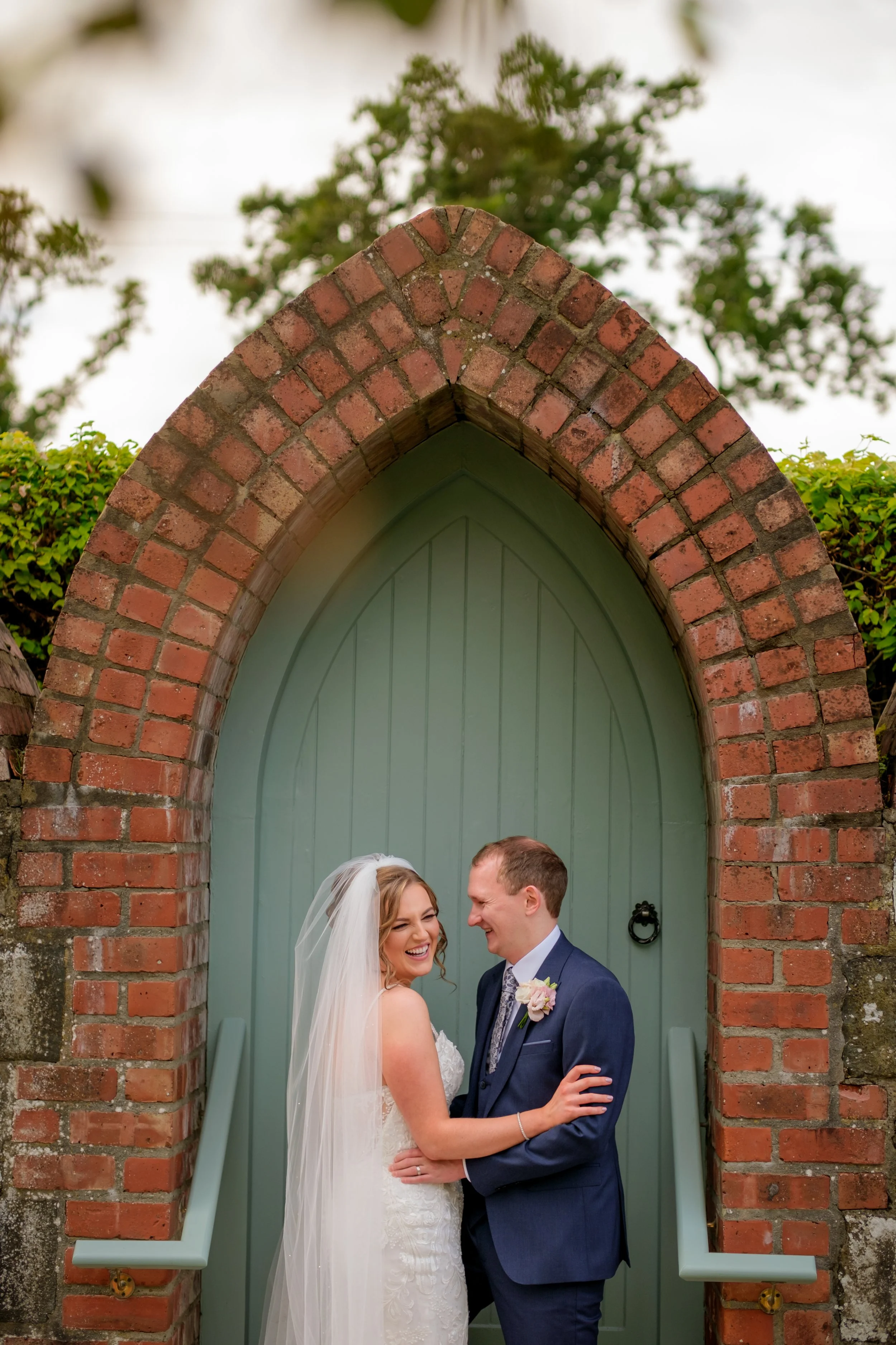 A bride and groom sharing a joyful moment in front of a green arched door framed by red brick archway during their wedding.