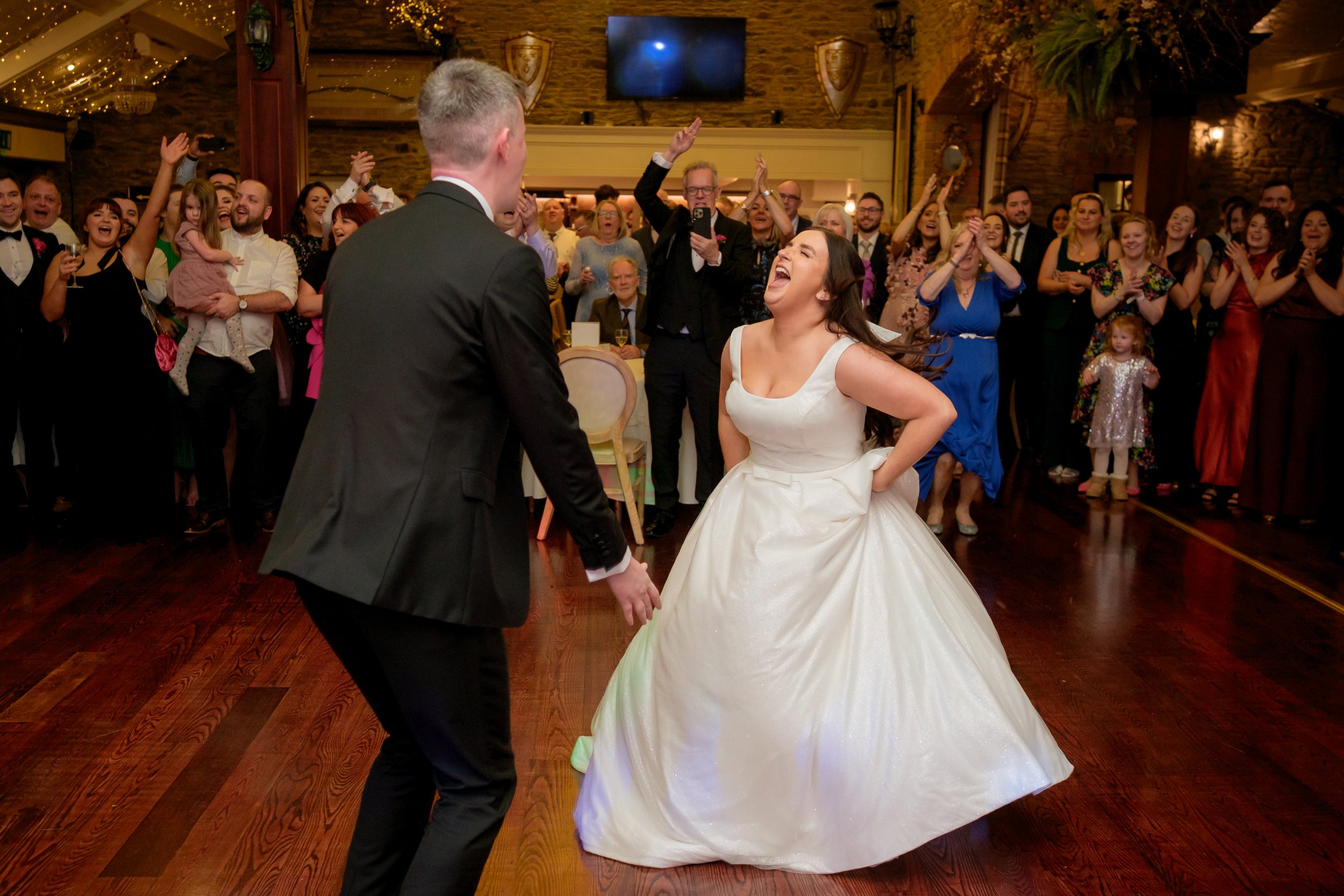 A bride and groom dancing at their wedding reception with guests clapping and cheering in the background.