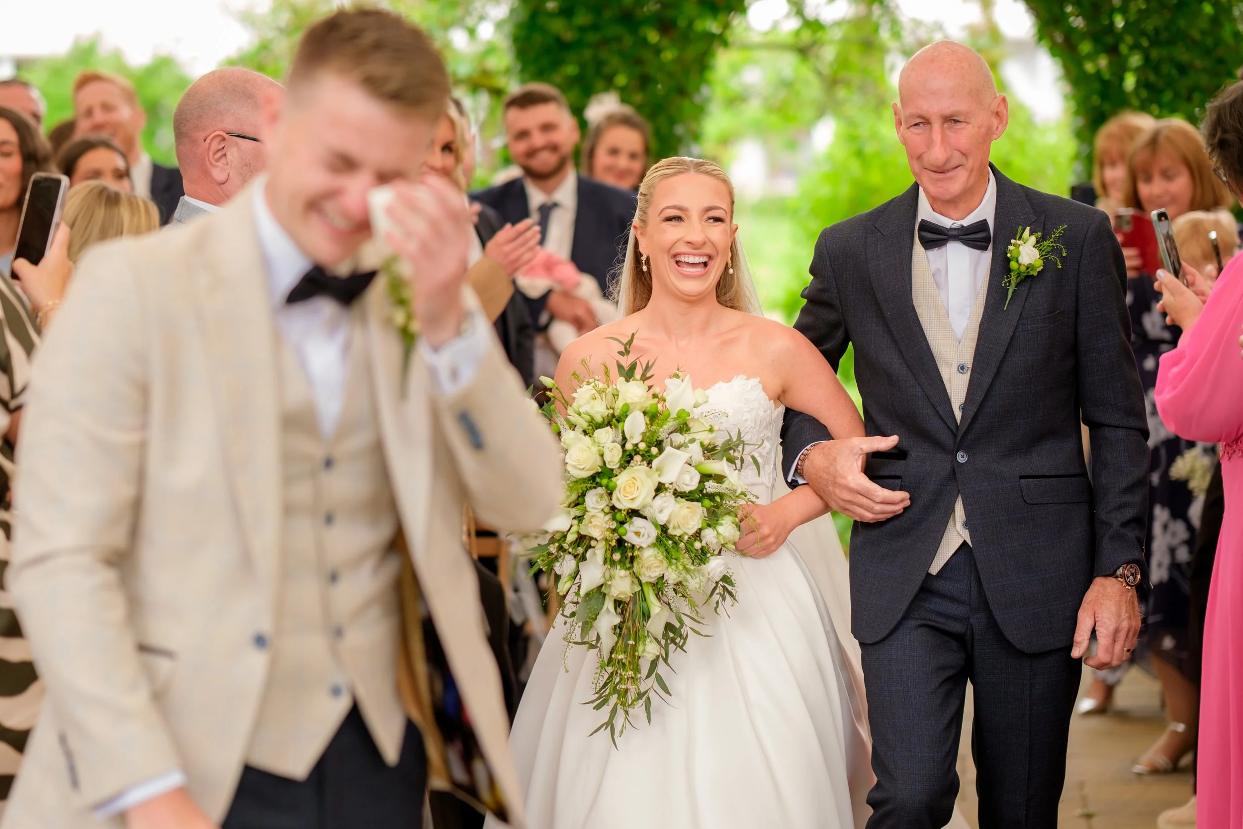 A bride smiling and holding a large white bouquet walking down the aisle with her father, surrounded by happy wedding guests. The groom appears emotional and is wiping tears from his face.