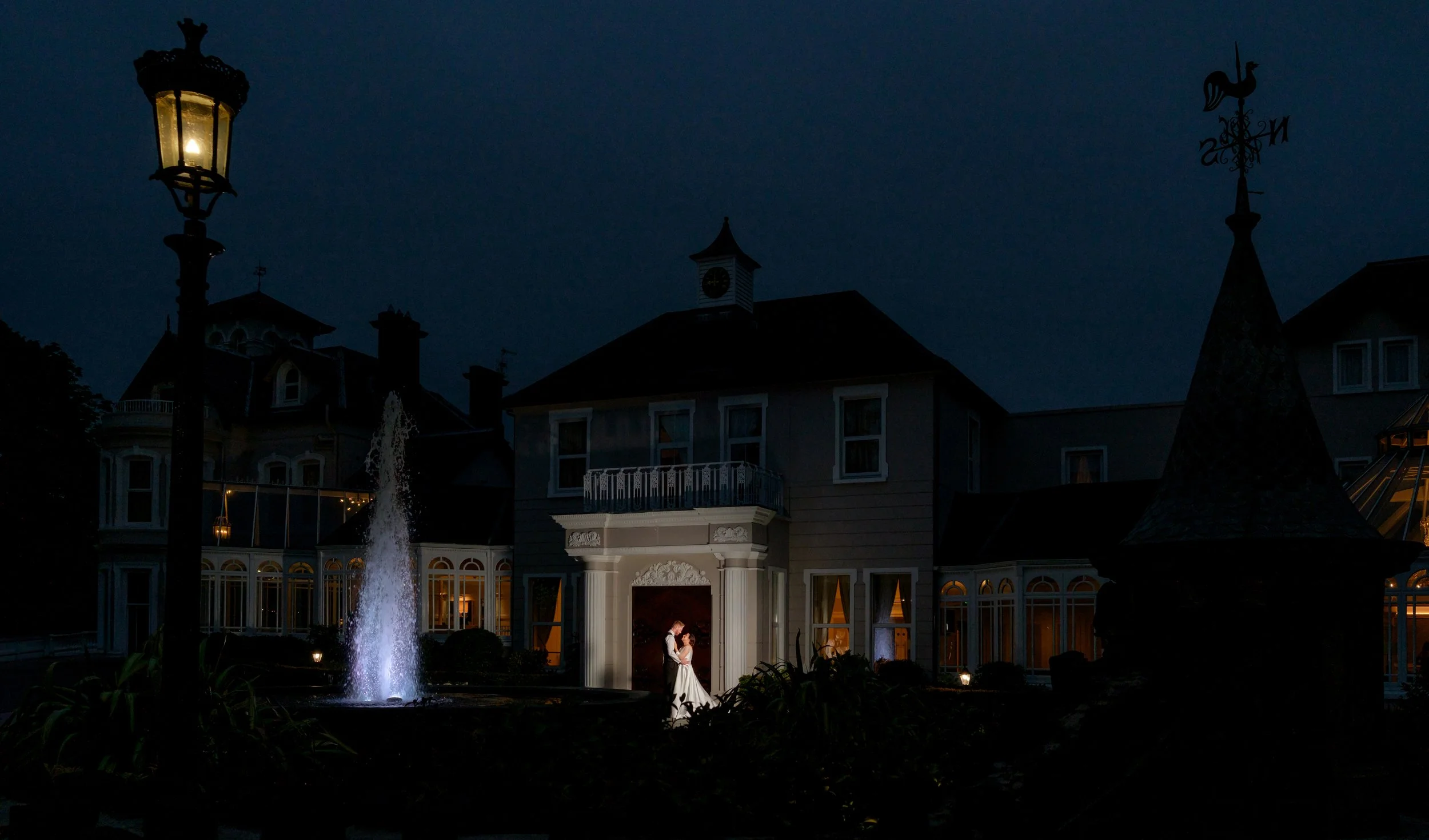 Nighttime scene of a large house with illuminated windows, a fountain, and a couple dancing under a porch light.