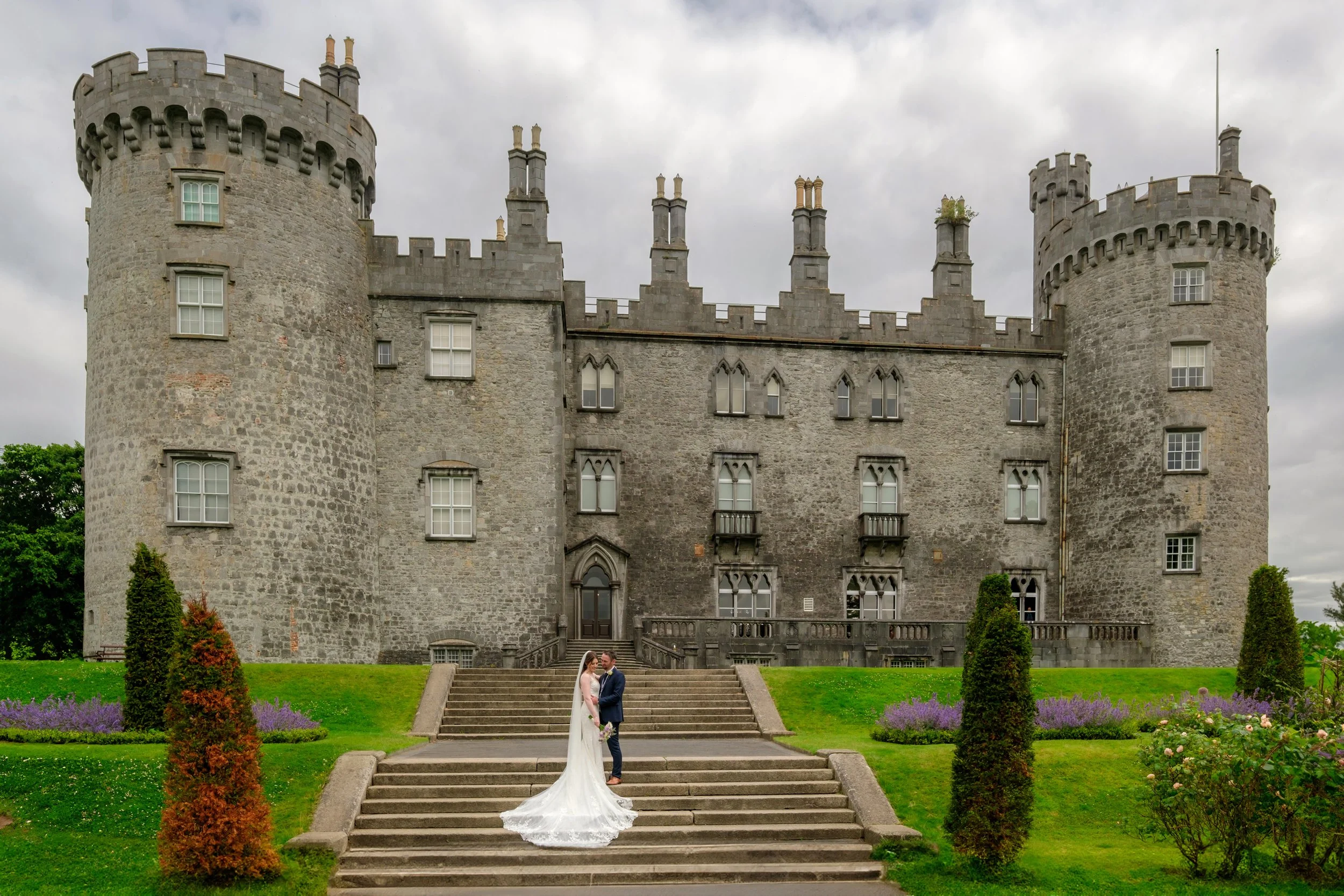 A bride and groom in wedding attire standing on stone stairs in front of a historic castle with turrets and stone walls, surrounded by manicured green lawns and flower bushes.