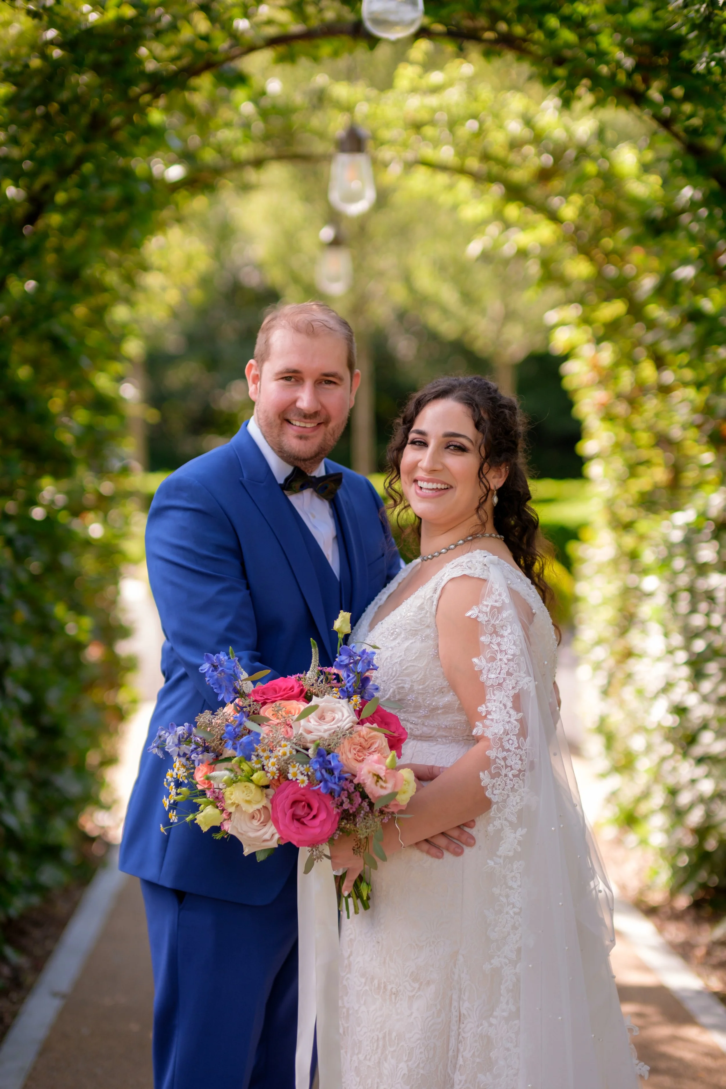 A newlywed couple standing outdoors on a garden path, smiling, with the groom in a blue suit and bow tie, holding a colorful bouquet of flowers, and the bride in a lace wedding dress with a bouquet, under a green archway with hanging lanterns.