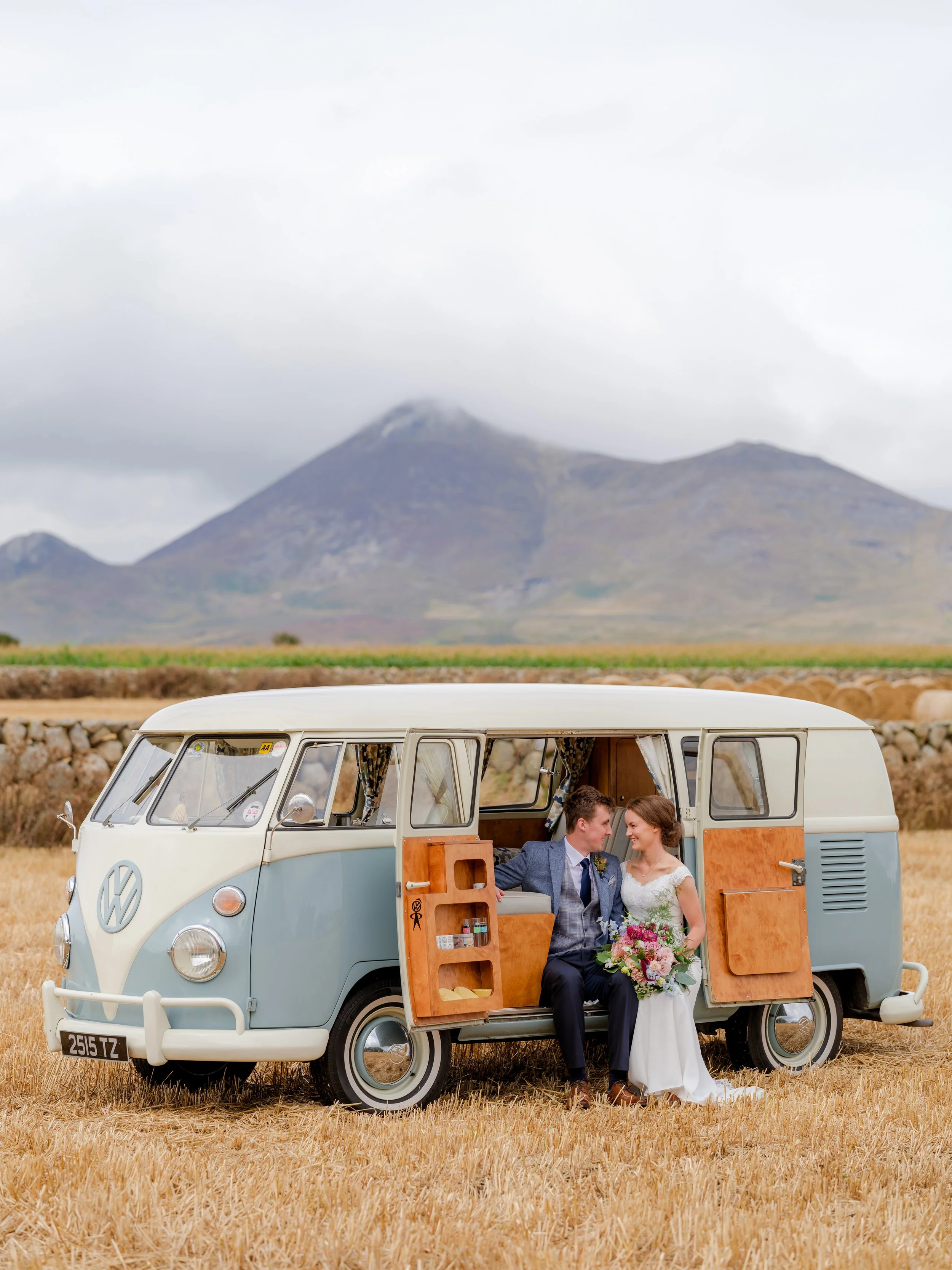 A couple in wedding attire sitting in a vintage Volkswagen camper van in a field, with mountains in the background.
