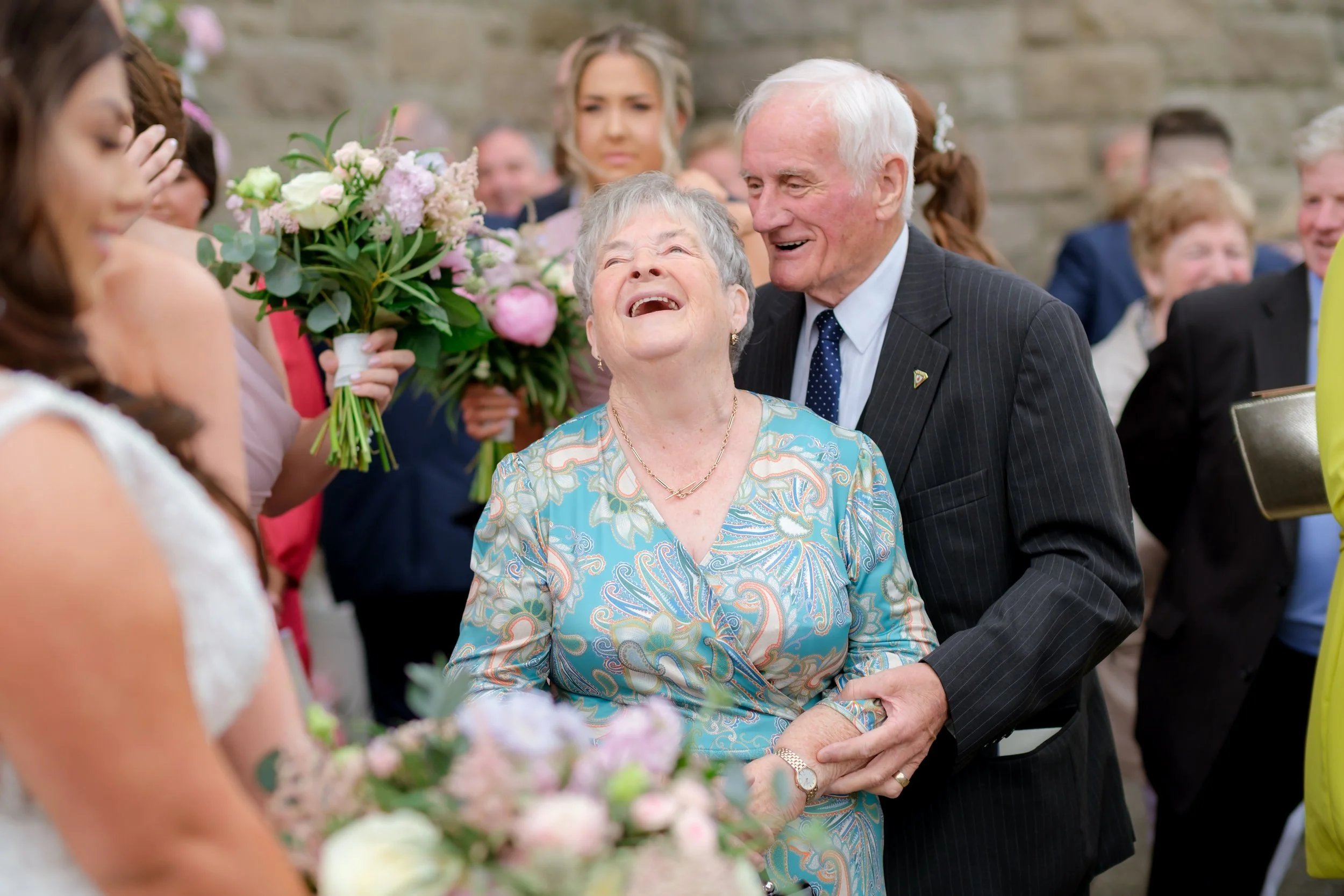 An elderly woman is laughing happily while holding hands with an elderly man at a wedding reception. The woman is wearing a colorful patterned dress, and the man is in a dark suit with a blue tie. Several women holding bouquets and other guests are i