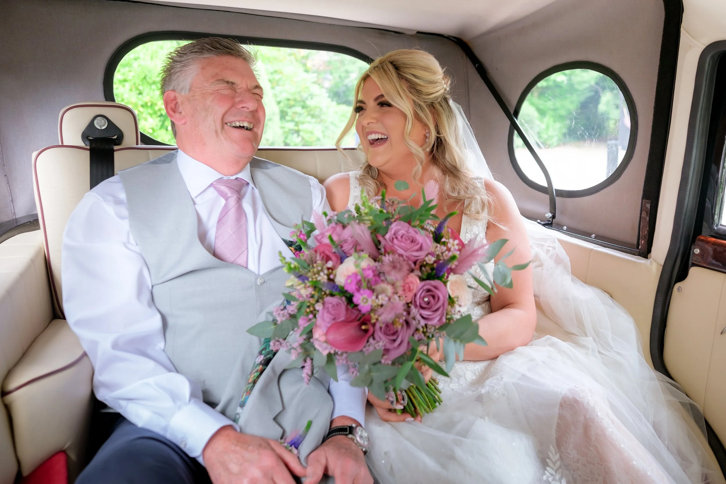 A bride and an older man, possibly her father, sitting inside a vehicle, both laughing happily. The bride is holding a large pink and purple bouquet and wearing a wedding dress, while the man is dressed in formal attire with a light gray vest and pin