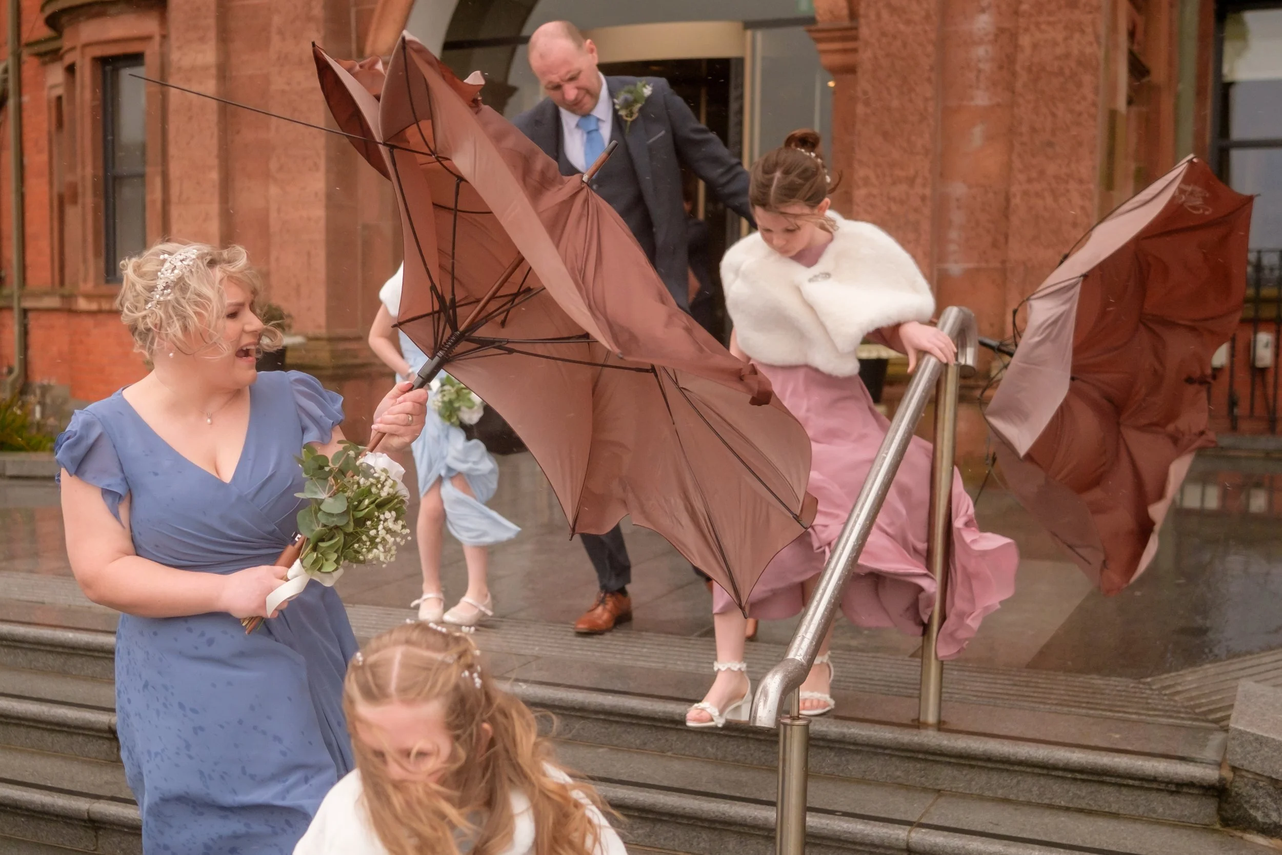 People in formal attire navigating wet stairs with umbrellas outside a brick building.