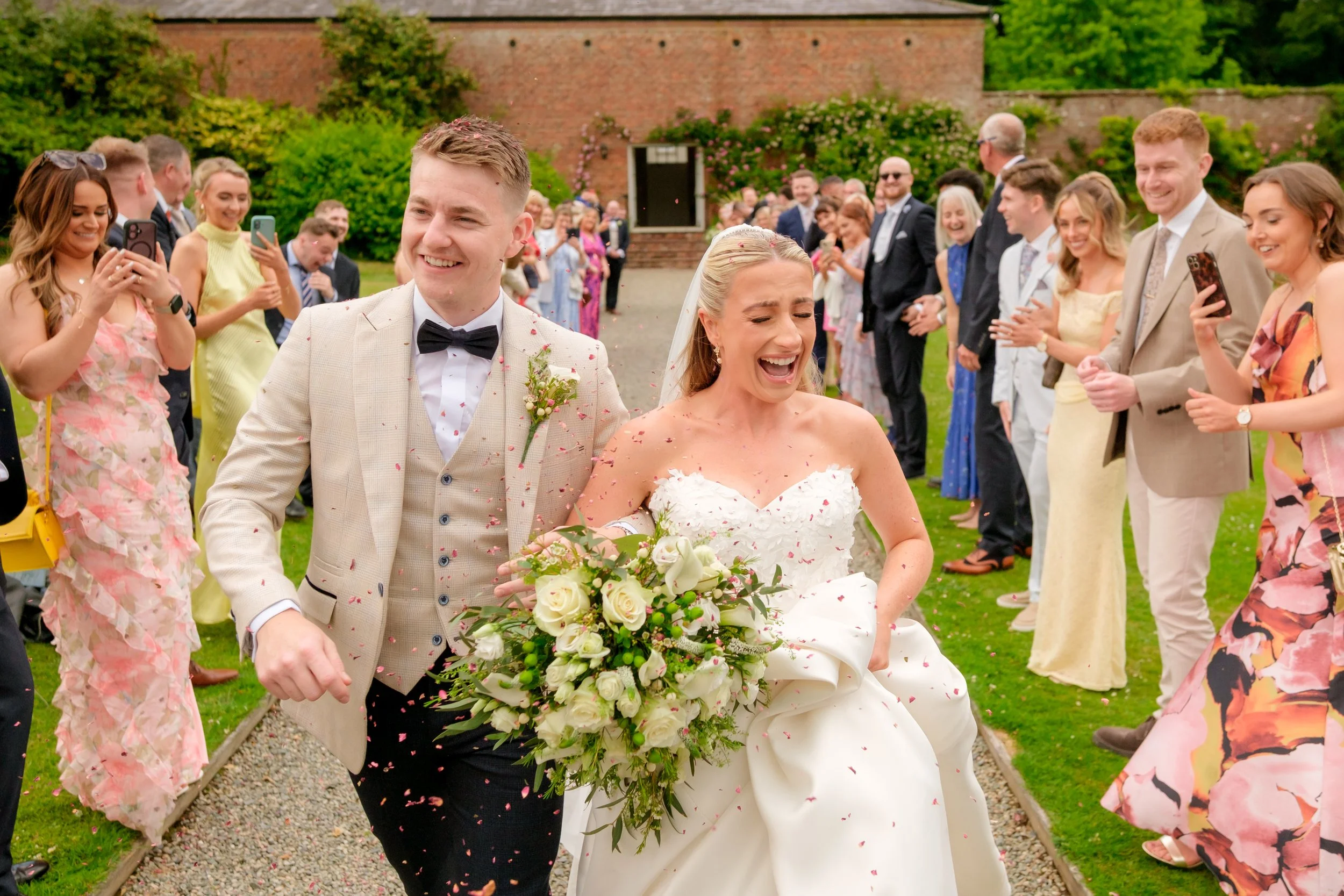Wedding celebration with bride and groom smiling and walking through crowd of guests throwing flower petals outdoors.