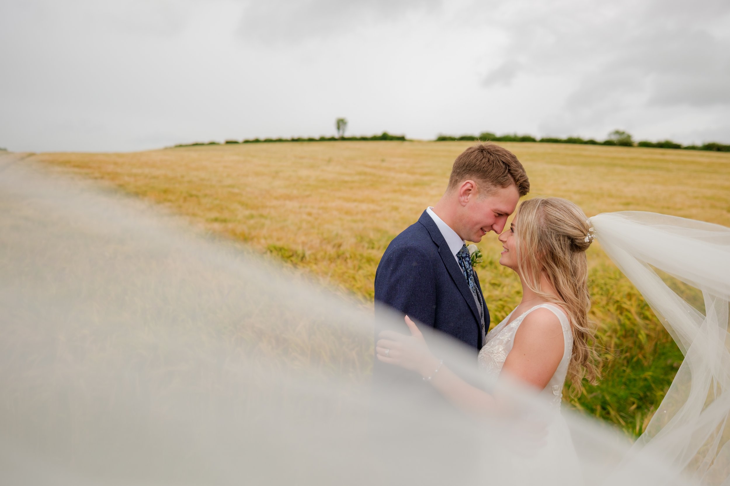 A bride and groom standing close together in a field on a cloudy day, touching foreheads and smiling