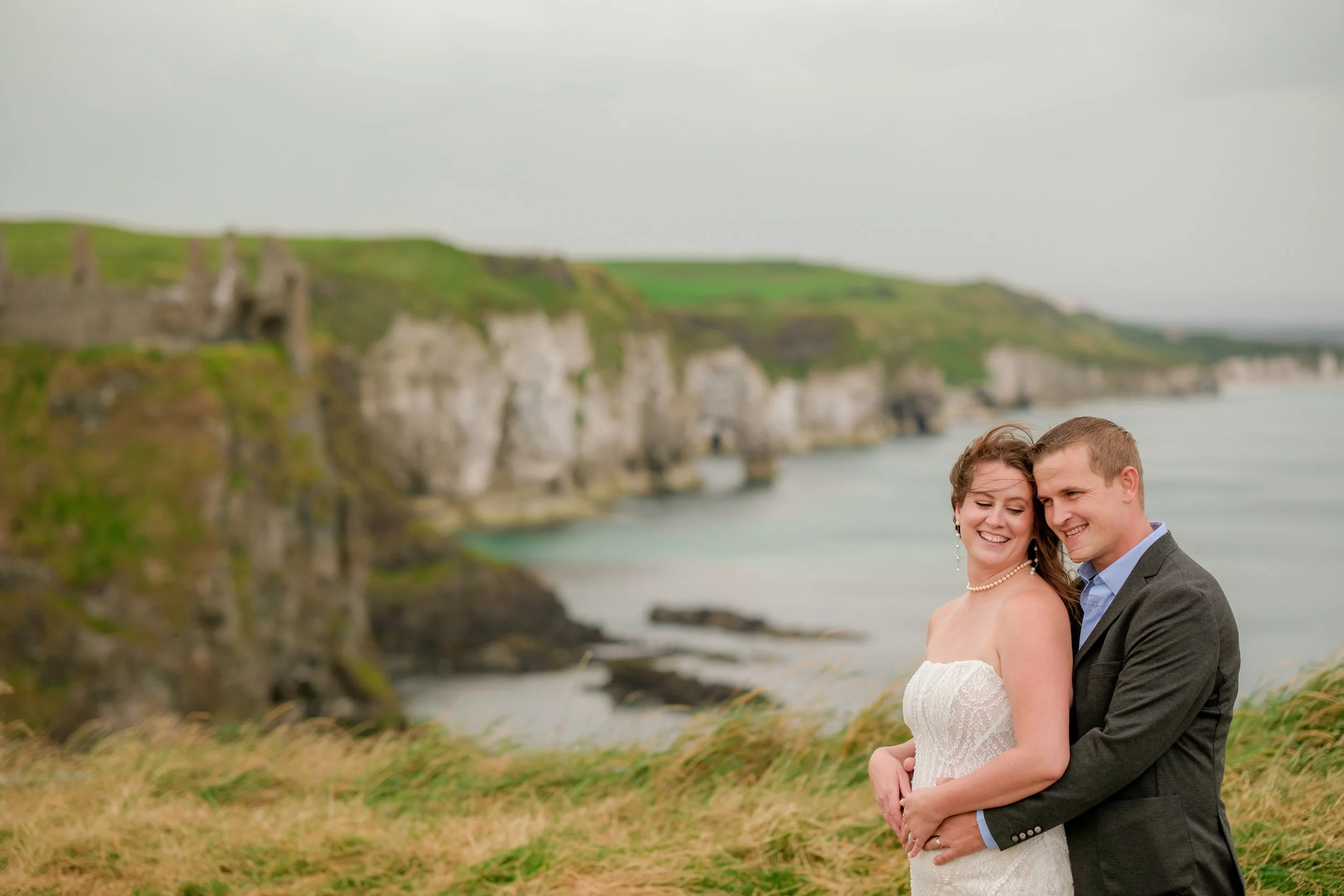 Couple in wedding attire embracing on a grassy hill with cliffs and ocean in the background.